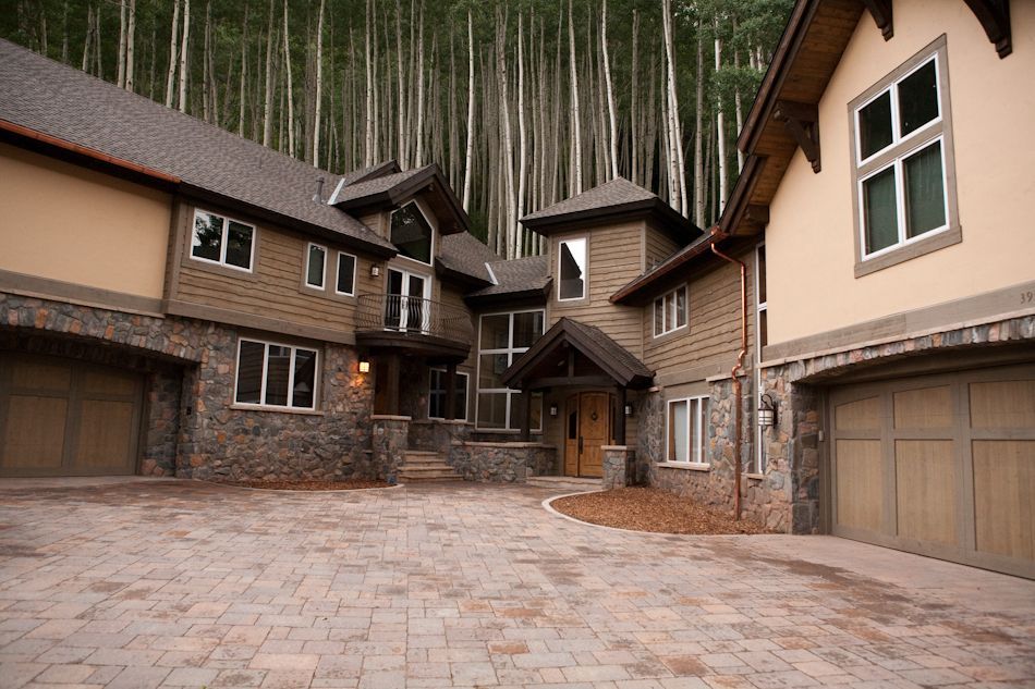 A large, multi-story mountain home with tan stucco, stone siding, and two garages, set against a forest of aspen trees.