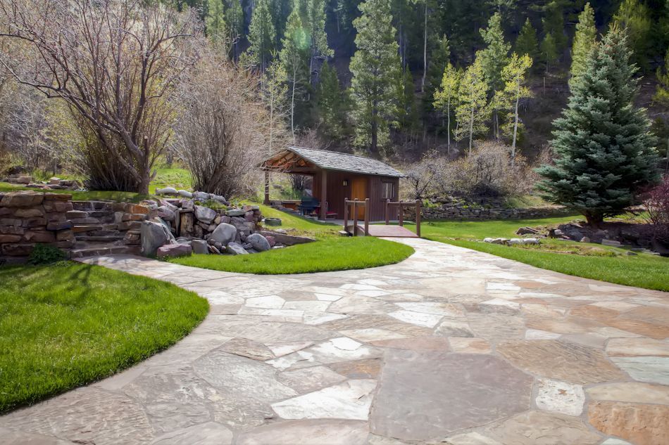 A stone patio leads to a wooden cabin nestled in a grassy, landscaped yard with trees and a rocky retaining wall.