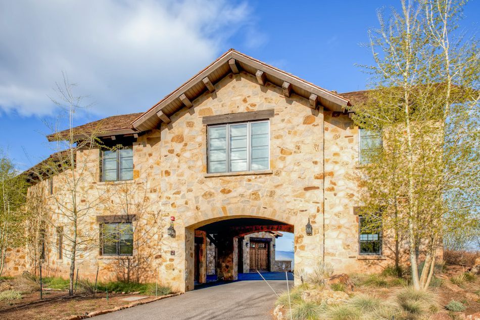 A two-story stone house with a central arched entryway and dark-framed windows, set against a blue sky with trees.