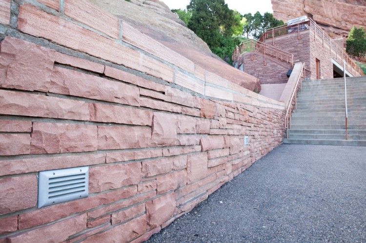 A red sandstone retaining wall with an inset light fixture next to stone stairs leading up to a rocky landscape.