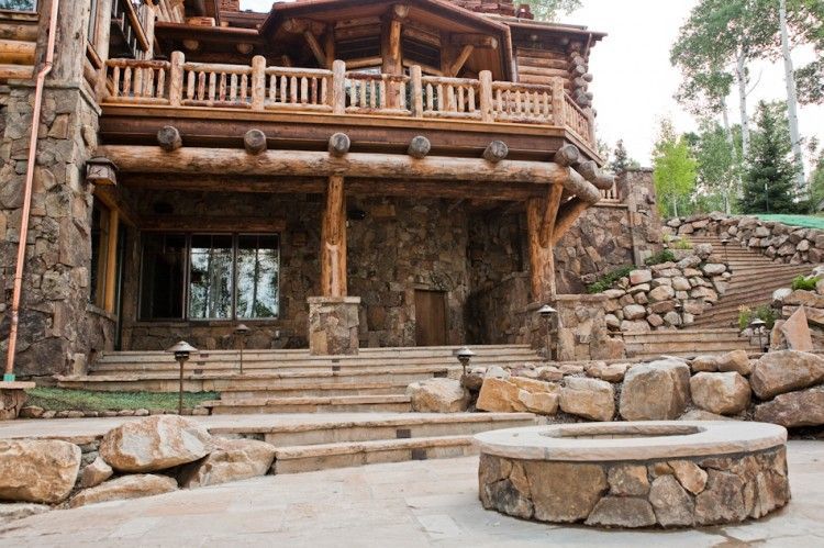A cozy stone courtyard with a metal gate, potted plants, a wooden barrel, and outdoor seating under a large autumn tree.