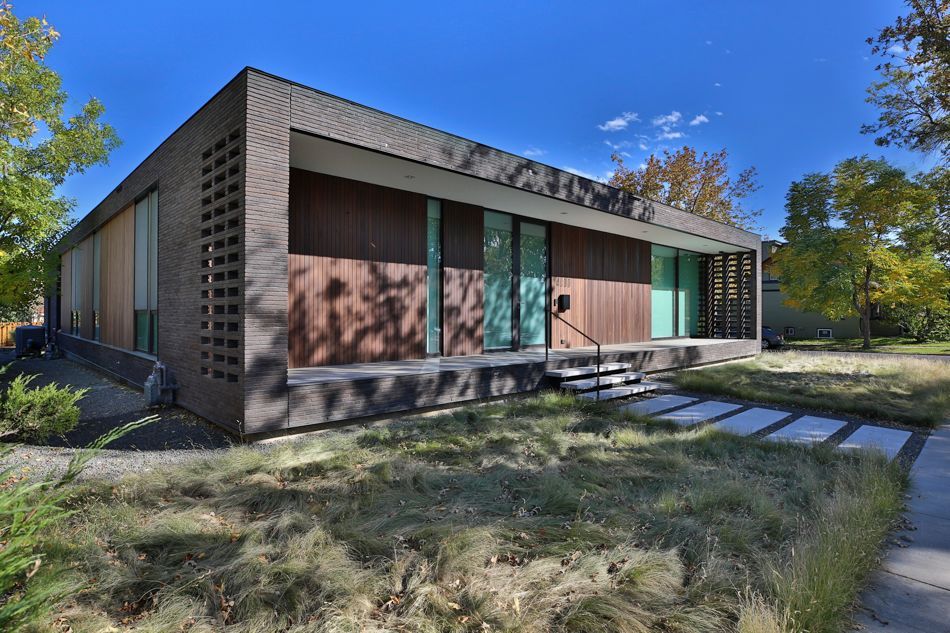 A contemporary single-story home with dark brick walls, wood paneling, and a paved walkway, surrounded by fall foliage.