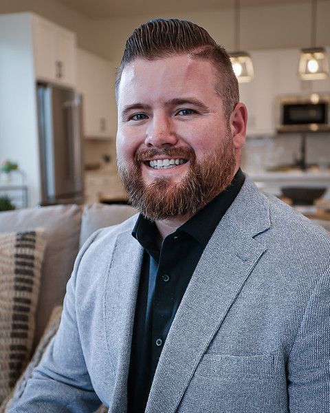 Man with beard smiling in a living room, wearing a gray blazer and black shirt.