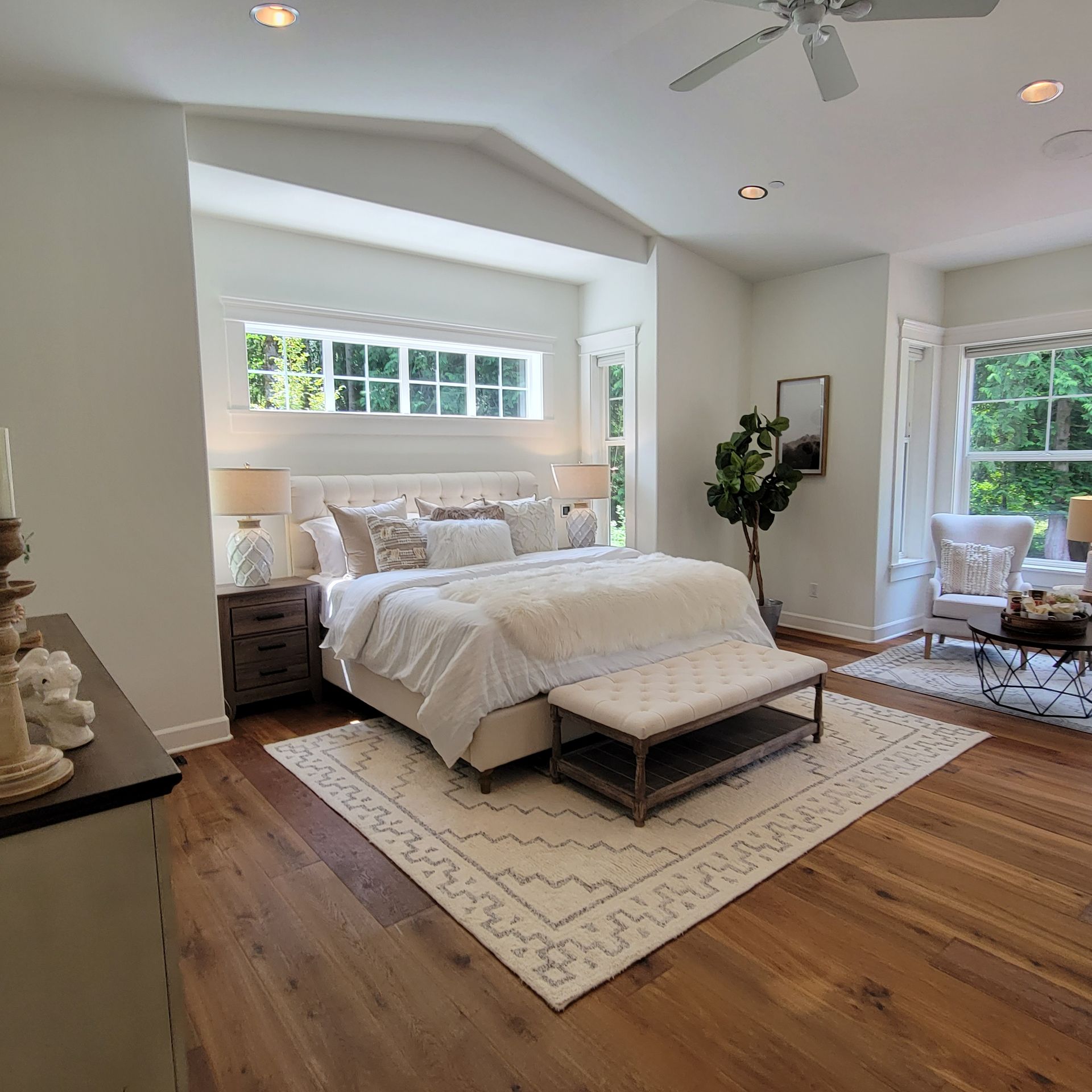 Cozy bedroom with a bed, rug, and sitting area near a window. Neutral tones with wooden floors and a ceiling fan.