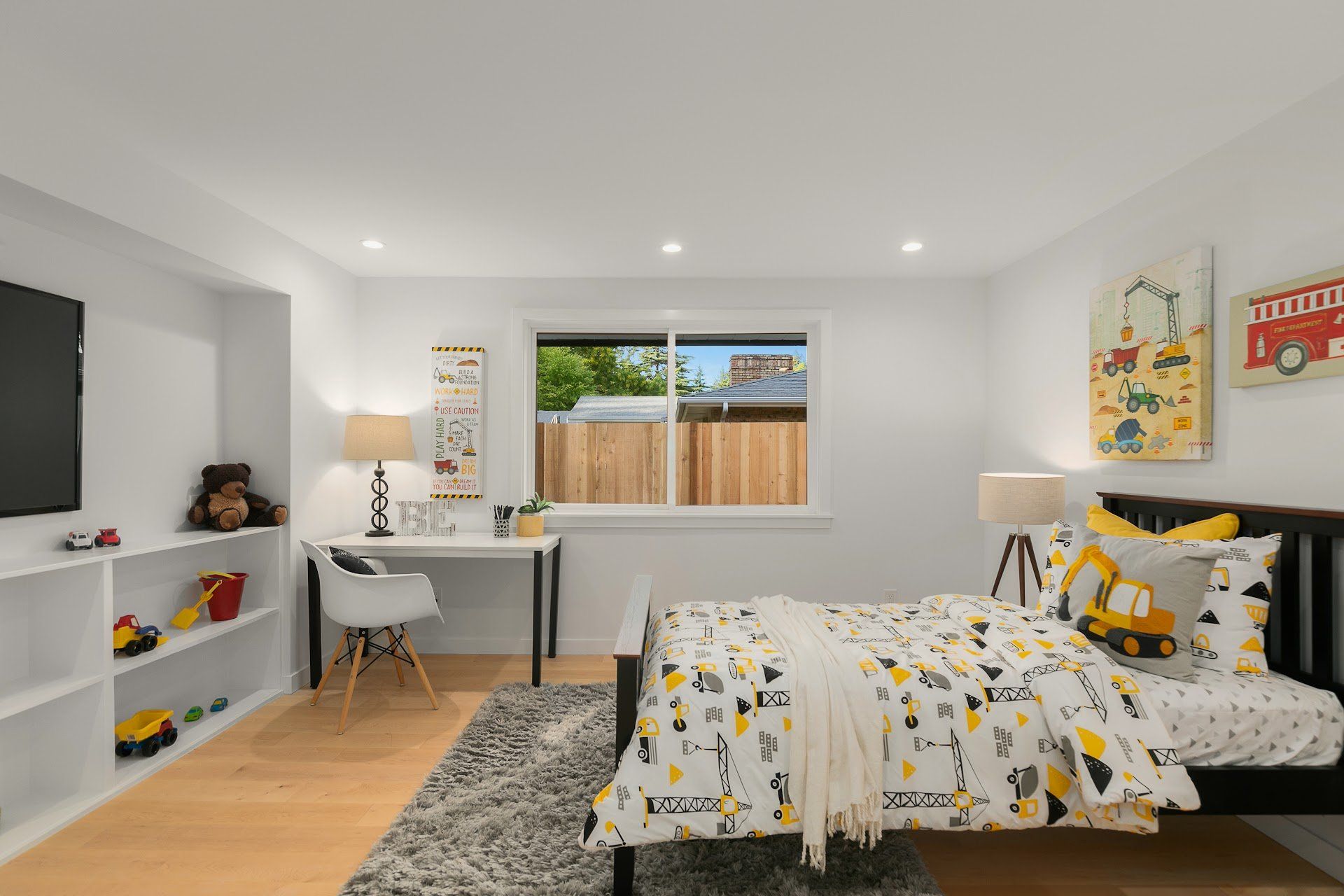 A child's bedroom with bed, desk, toys, and wall art featuring construction vehicles.