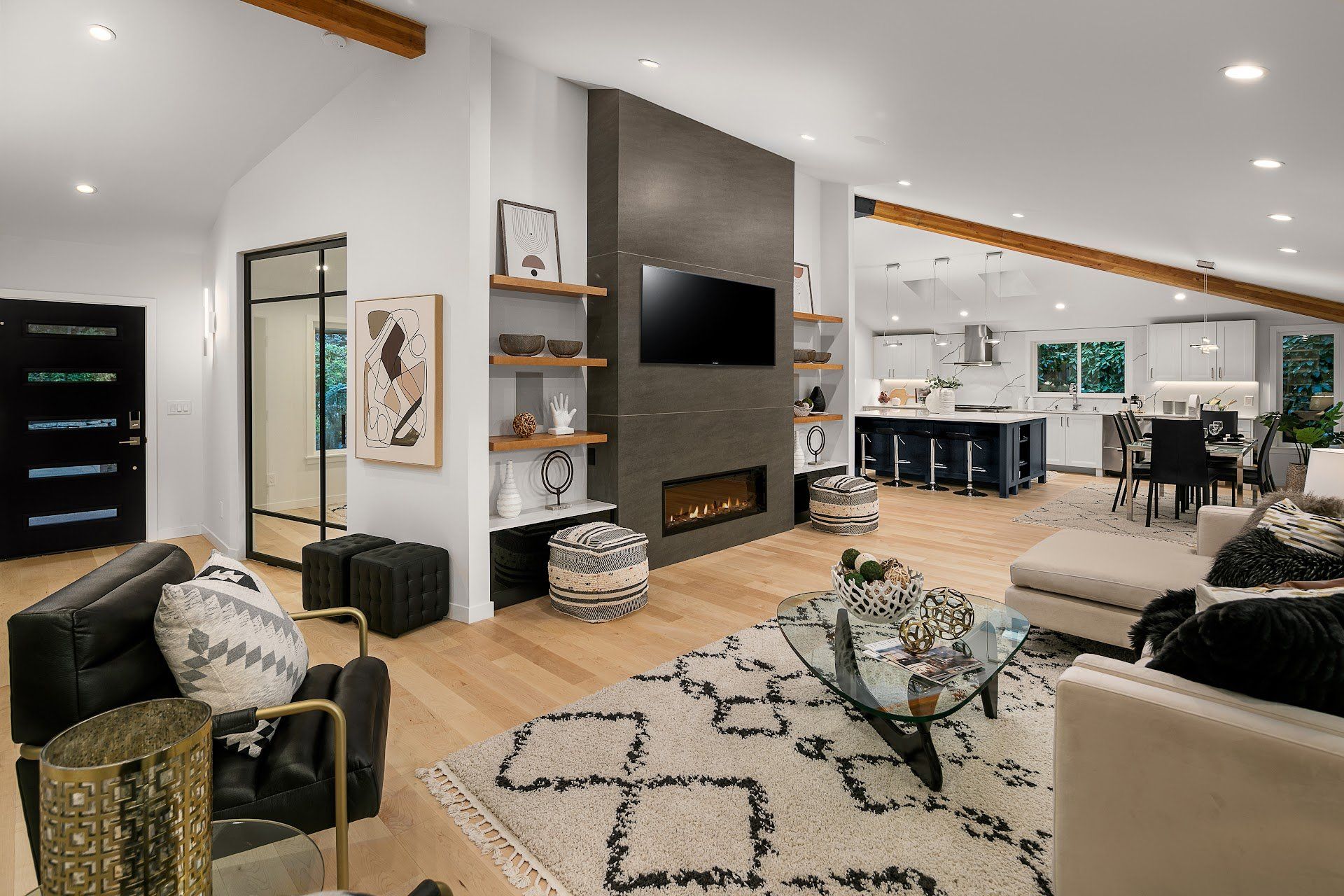 Modern living room with fireplace, TV, and open kitchen. Light wood floors, white walls, black and gray accents.