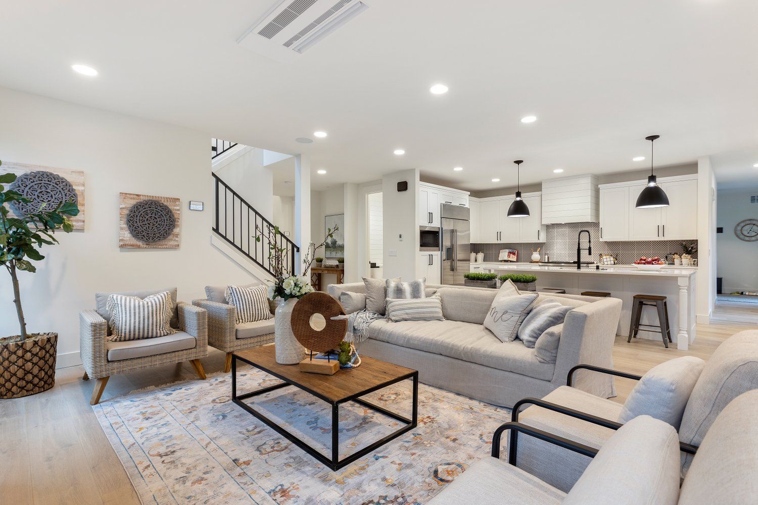 Bright, open-concept living space with kitchen, seating area, and staircase. White walls, light wood floors, and neutral furniture.