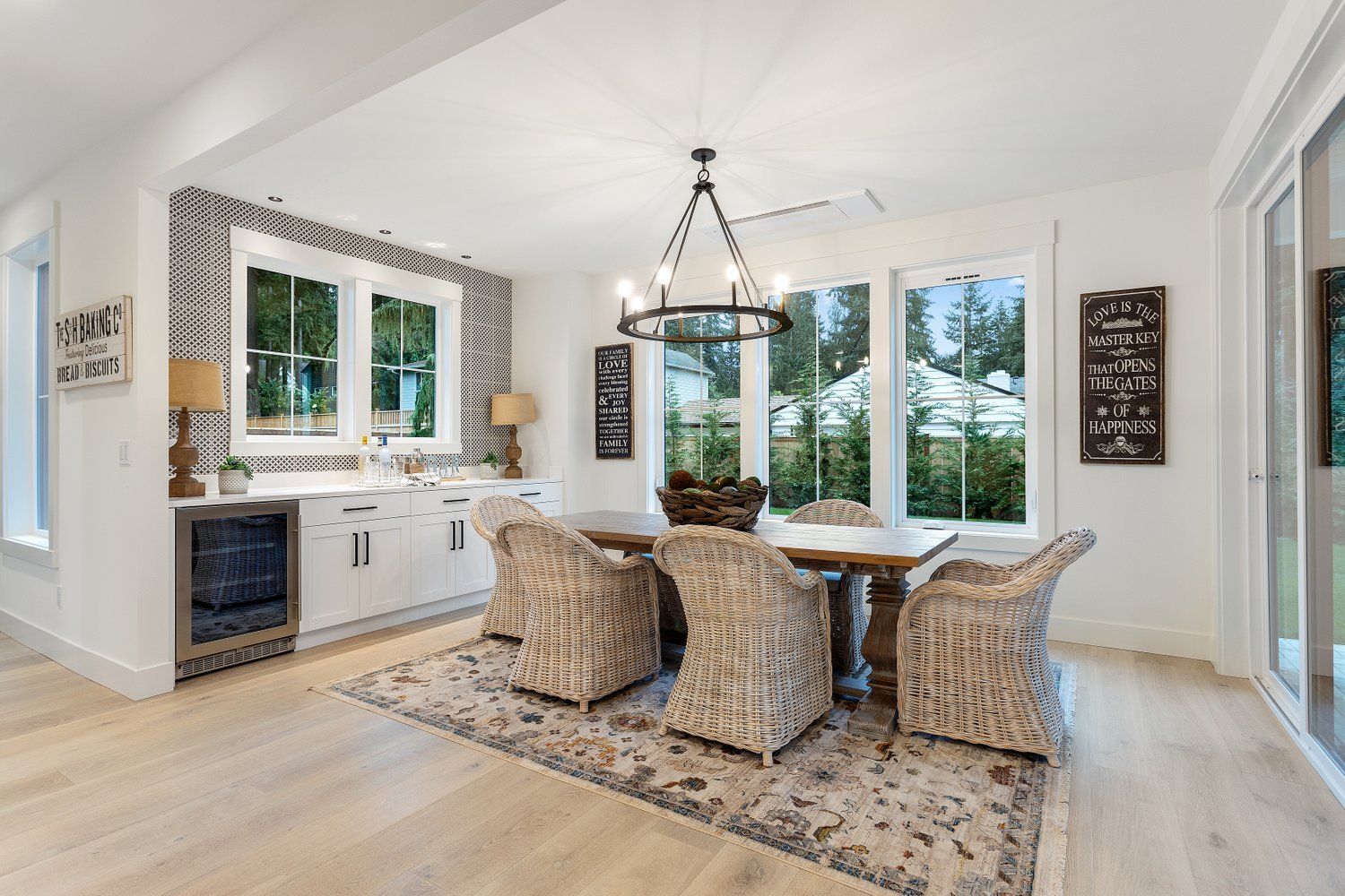 Bright dining room with wooden table, woven chairs, and large windows. White walls, patterned backsplash, and chandelier.