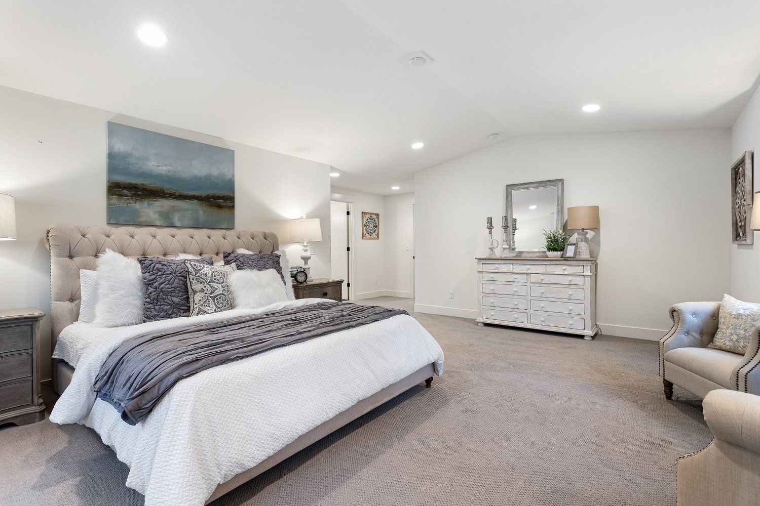 Spacious, bright bedroom with a tufted bed, dresser, armchair, and artwork; neutral tones.