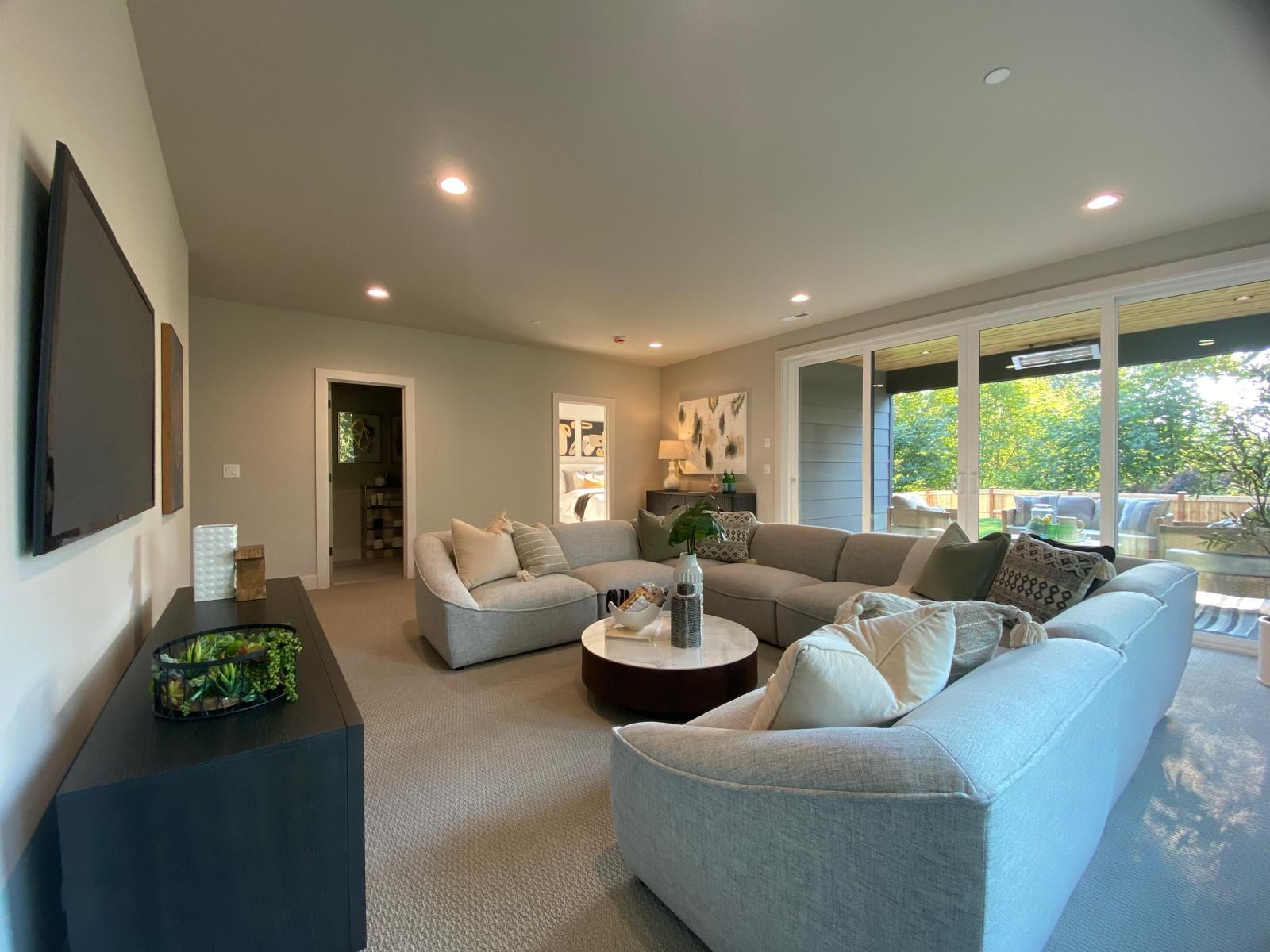 Modern living room with sectional sofa, coffee table, TV, and large windows overlooking a backyard.