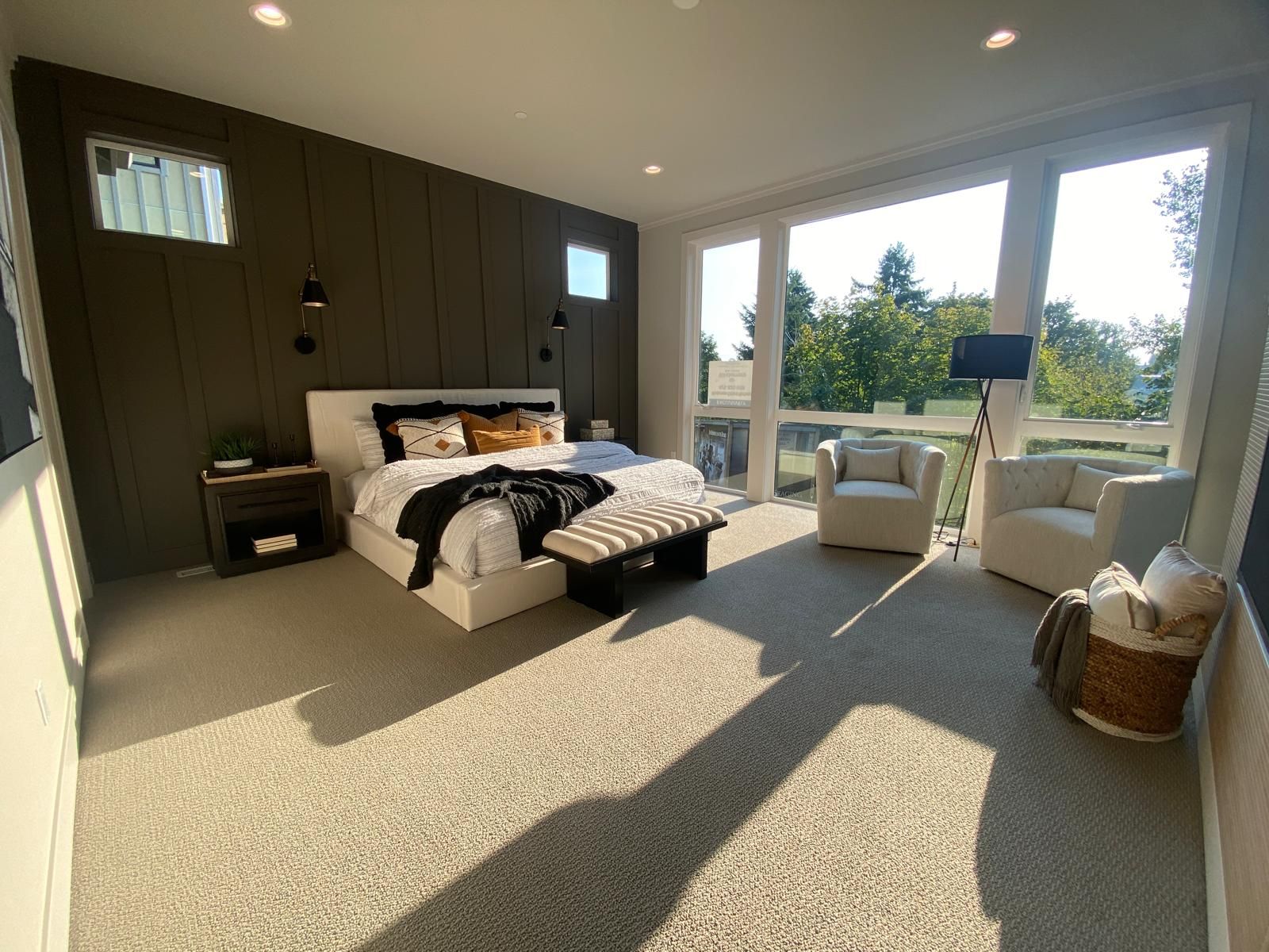 Spacious modern bedroom with a large window, bed, and seating area. Dark accent wall, neutral carpet.