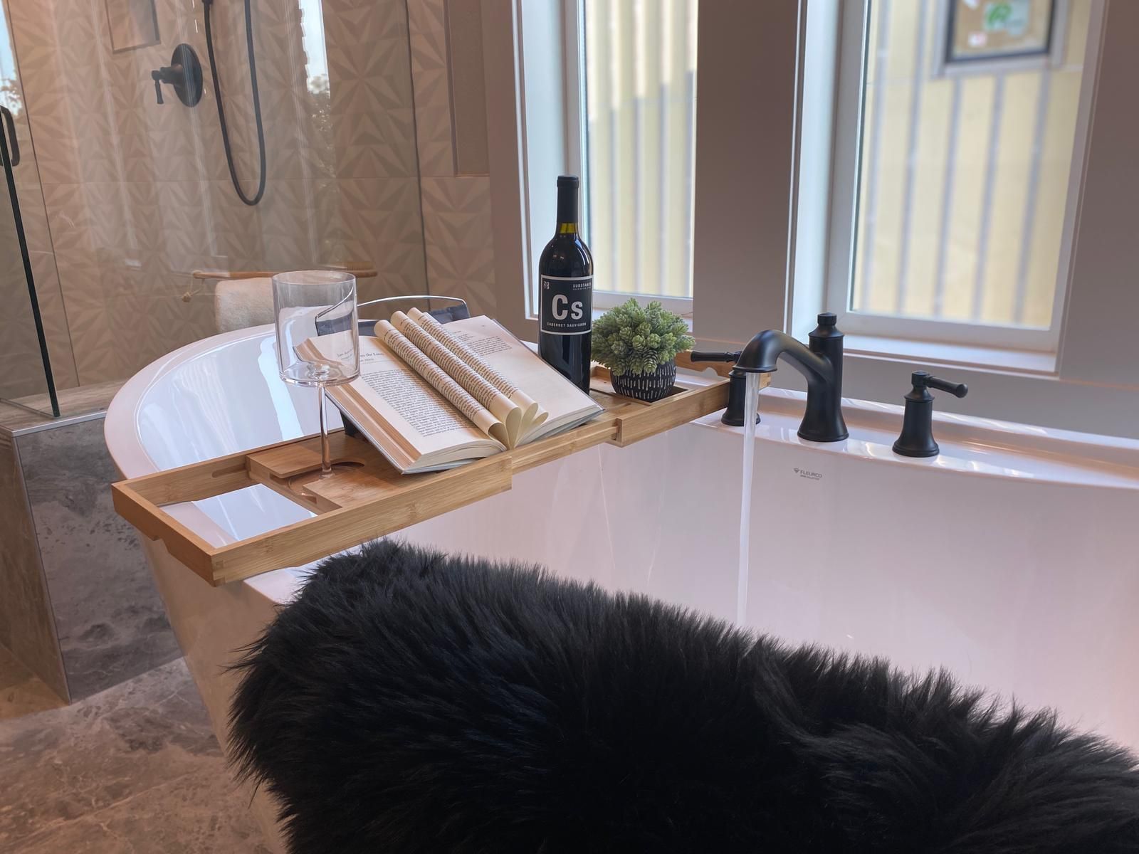 Bathtub with wooden tray holding wine, book, and plant; water running from faucet.