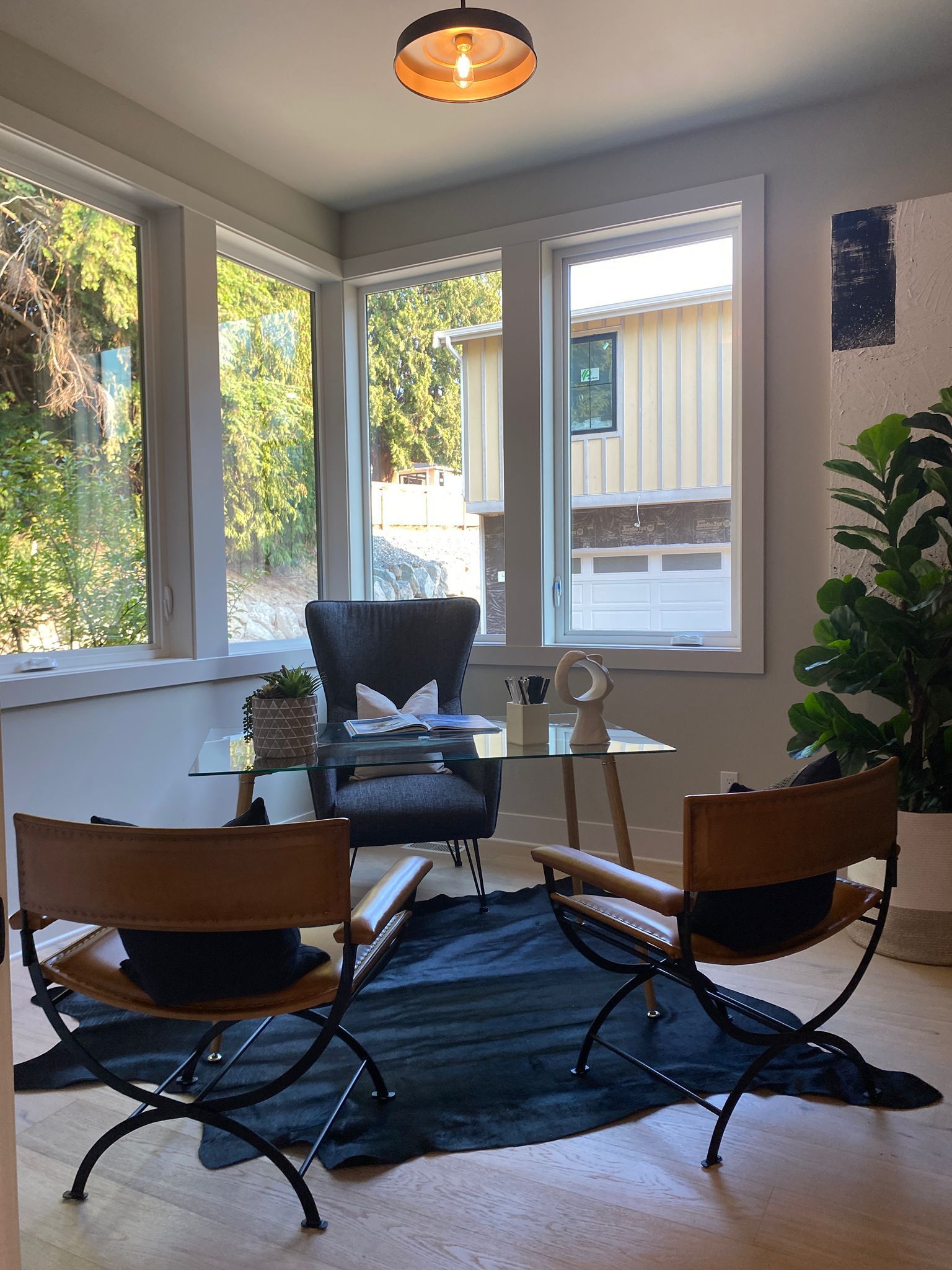 Office space with glass desk, leather chairs on a dark rug, windows overlooking trees and a house.