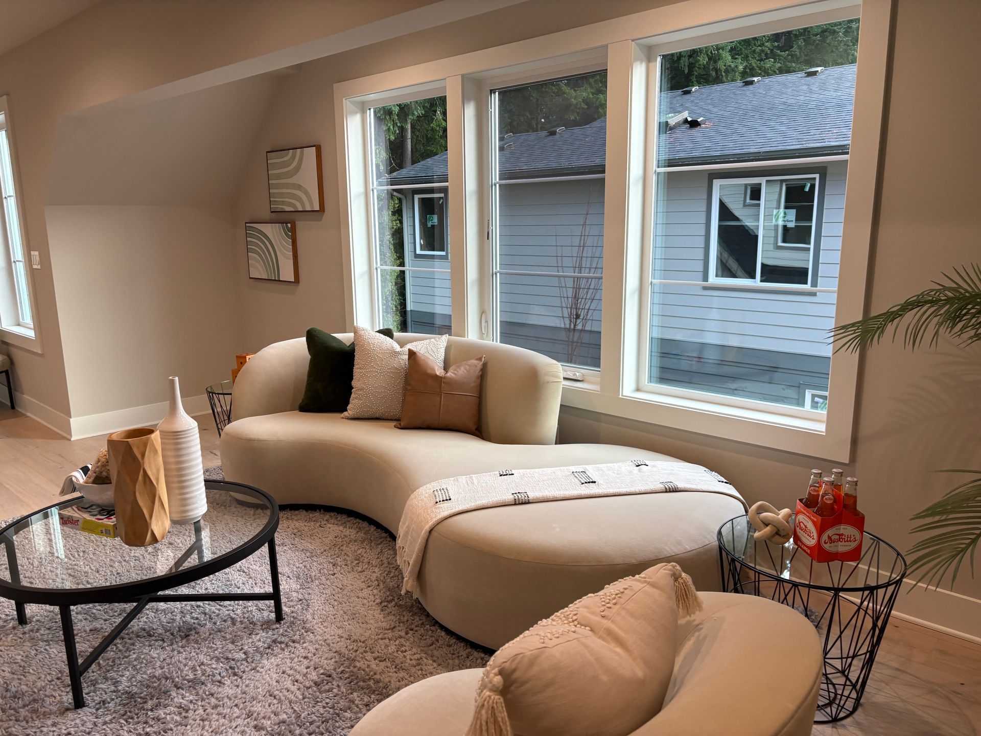 Living room with a curved sofa, round coffee table, and large windows overlooking a house.