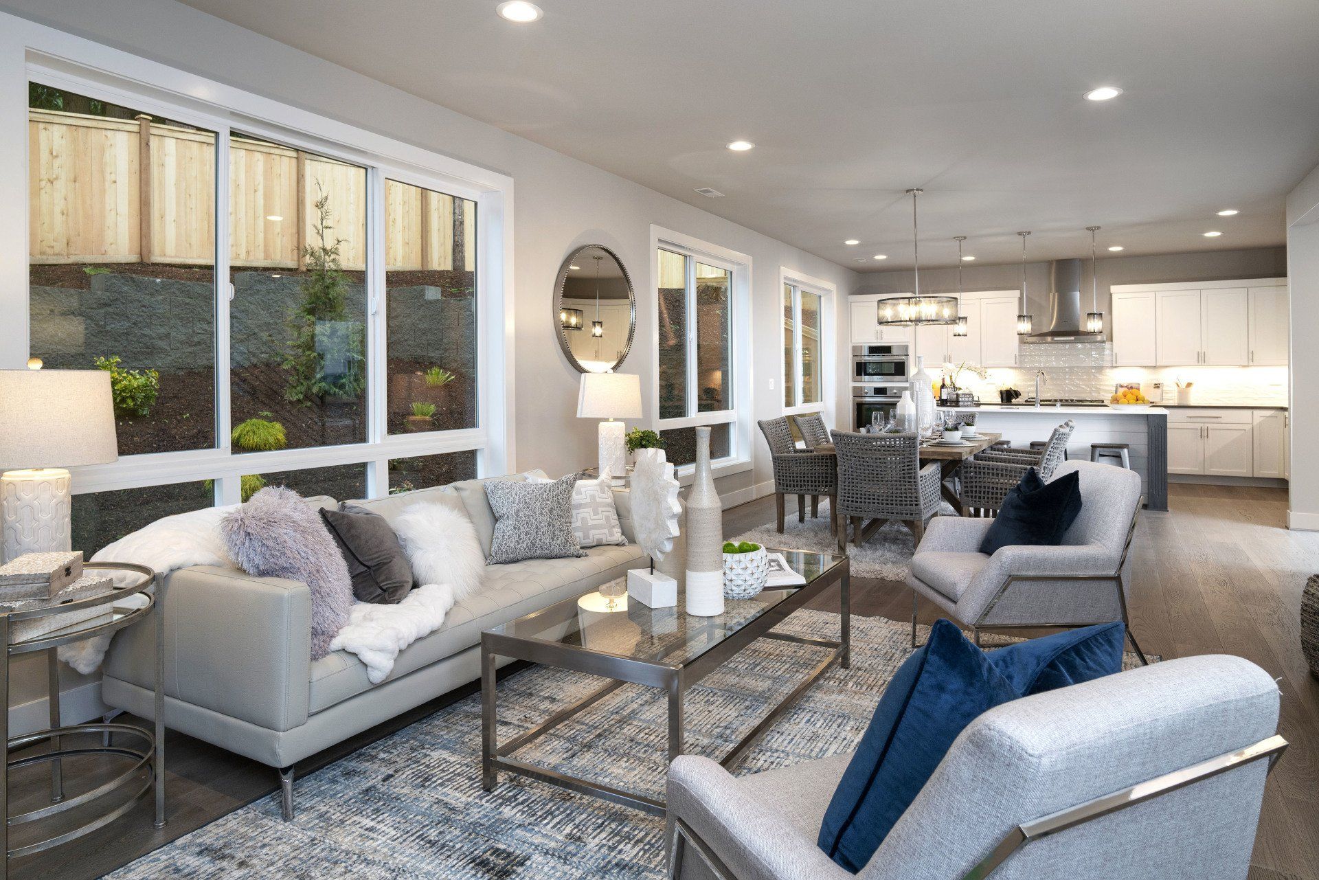 Modern living room with gray sofa, silver chairs, large windows, and open kitchen.