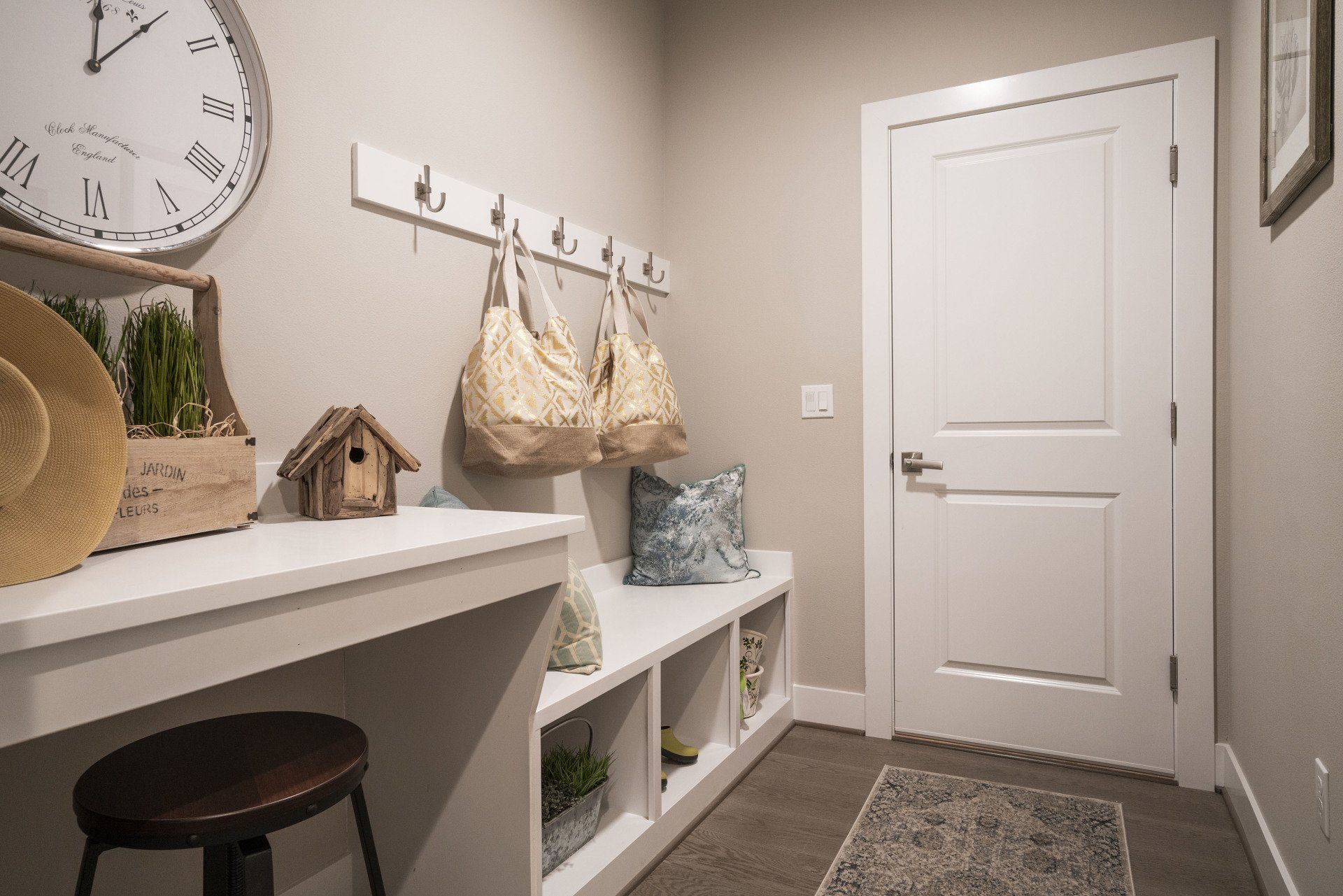 Mudroom with a white bench, hooks, and door; a large clock and stool sit beside a desk.