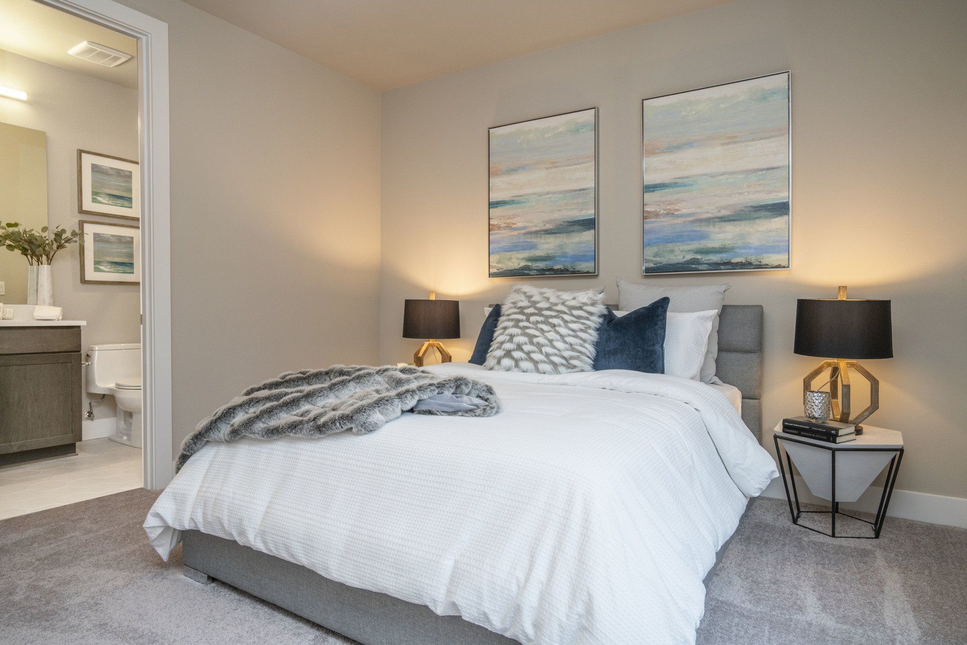 Bedroom with a bed, artwork, nightstands, and a bathroom visible. Light gray walls and carpet.