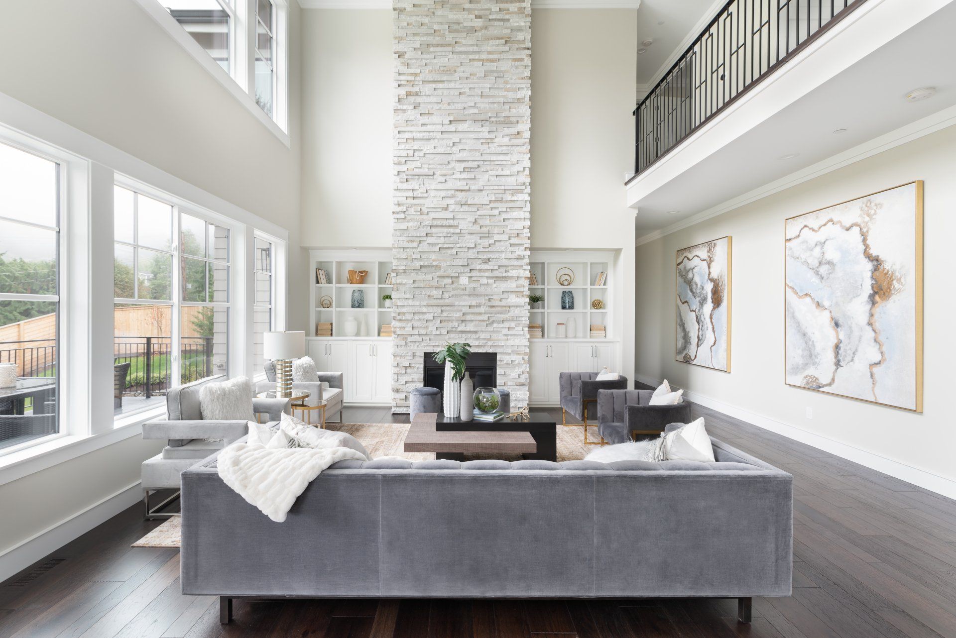 Spacious living room with high ceilings, large windows, fireplace, gray furniture, and hardwood floors.