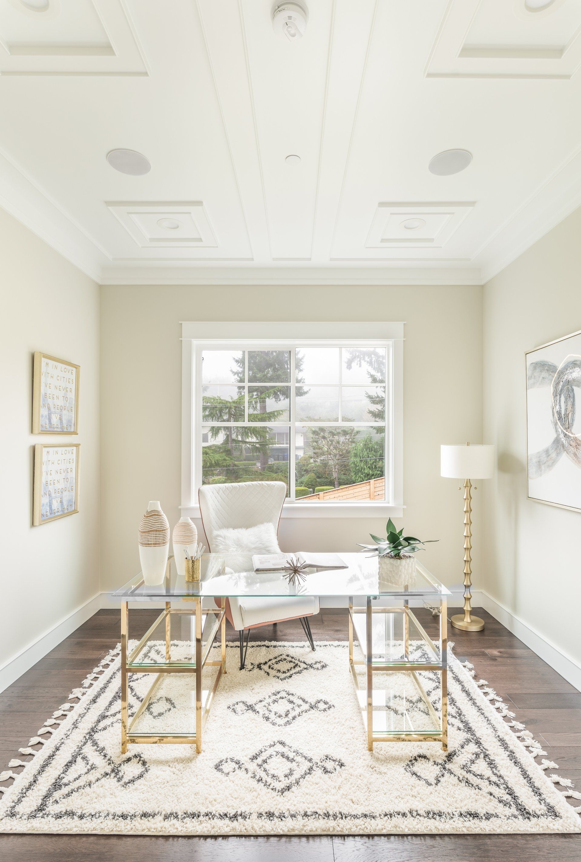 Cozy home office with a glass desk, gold accents, cream rug, and a window overlooking trees.