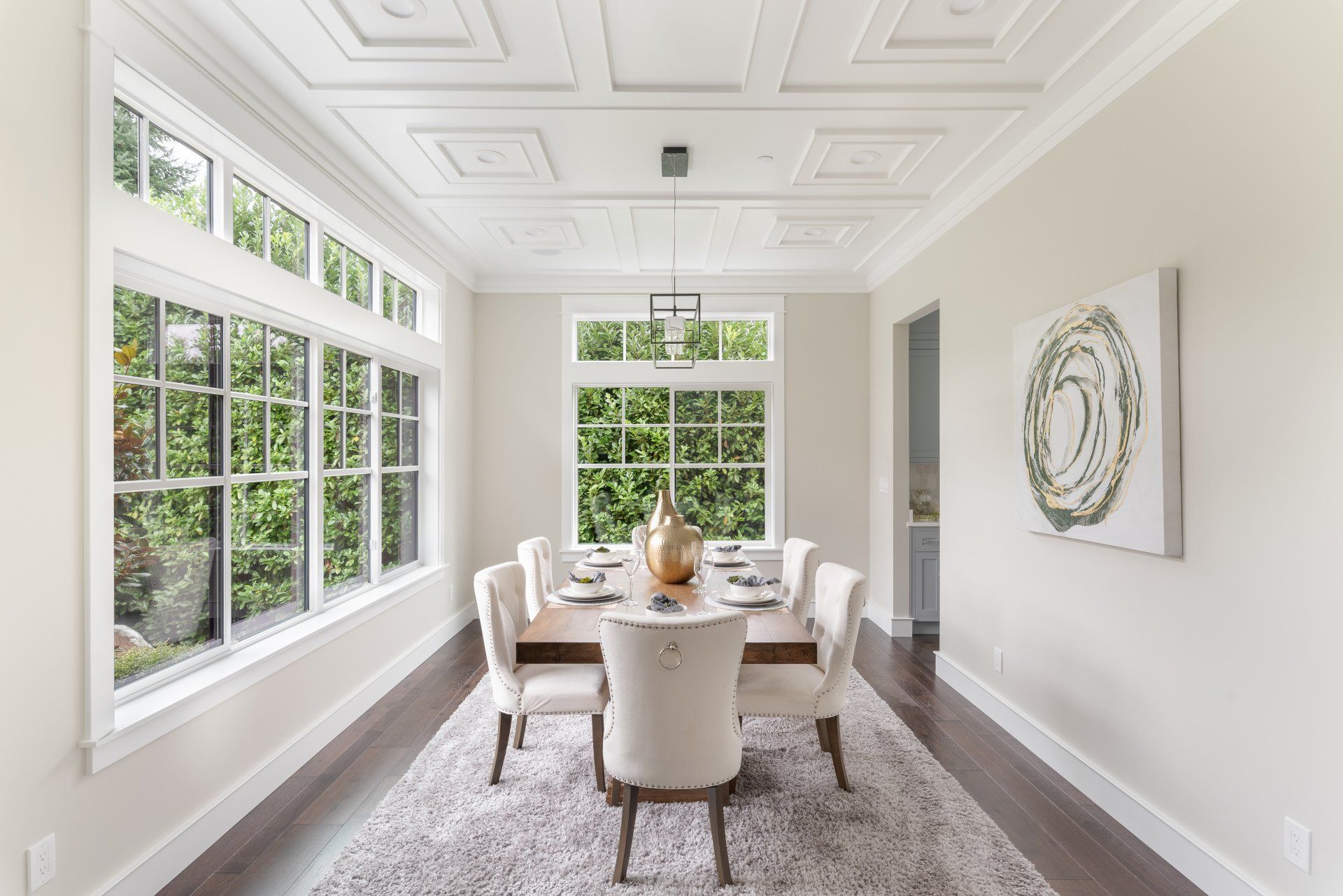 Dining room with a wooden table set for dinner, surrounded by cream chairs, with large windows and a patterned rug.