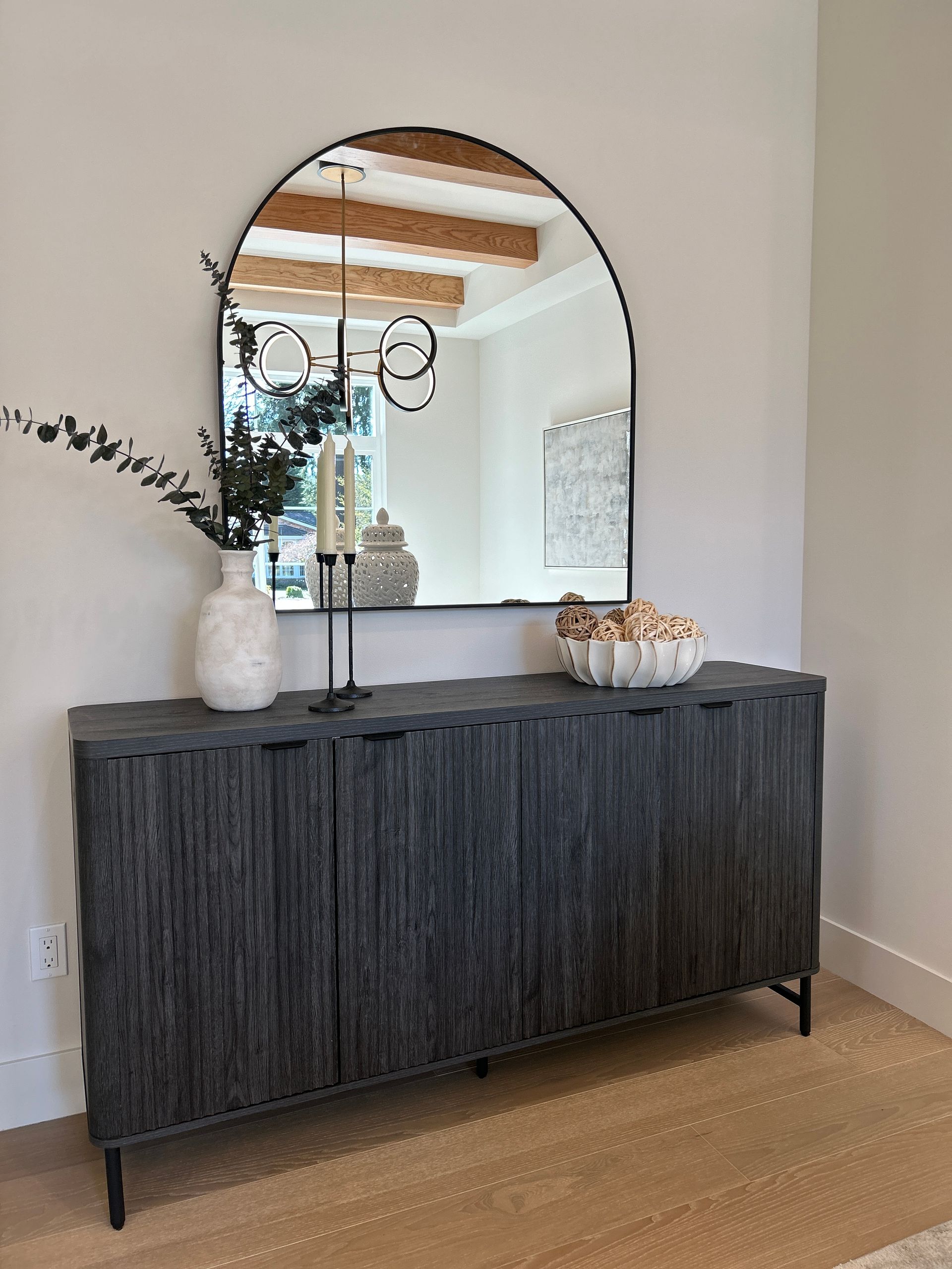 Dark wood sideboard beneath an arched mirror in a bright room, with a vase, candleholder, and fruit bowl.