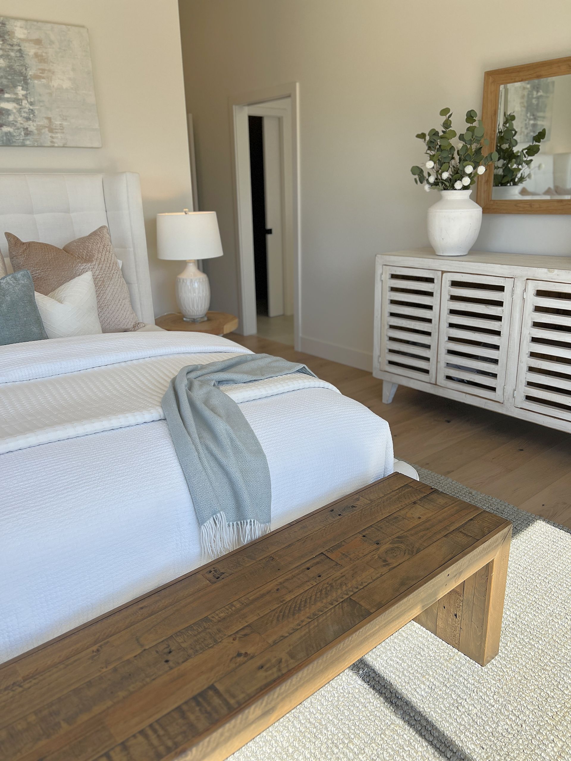 Bright bedroom with white bed, wooden bench, and slatted cabinet with vase and mirror