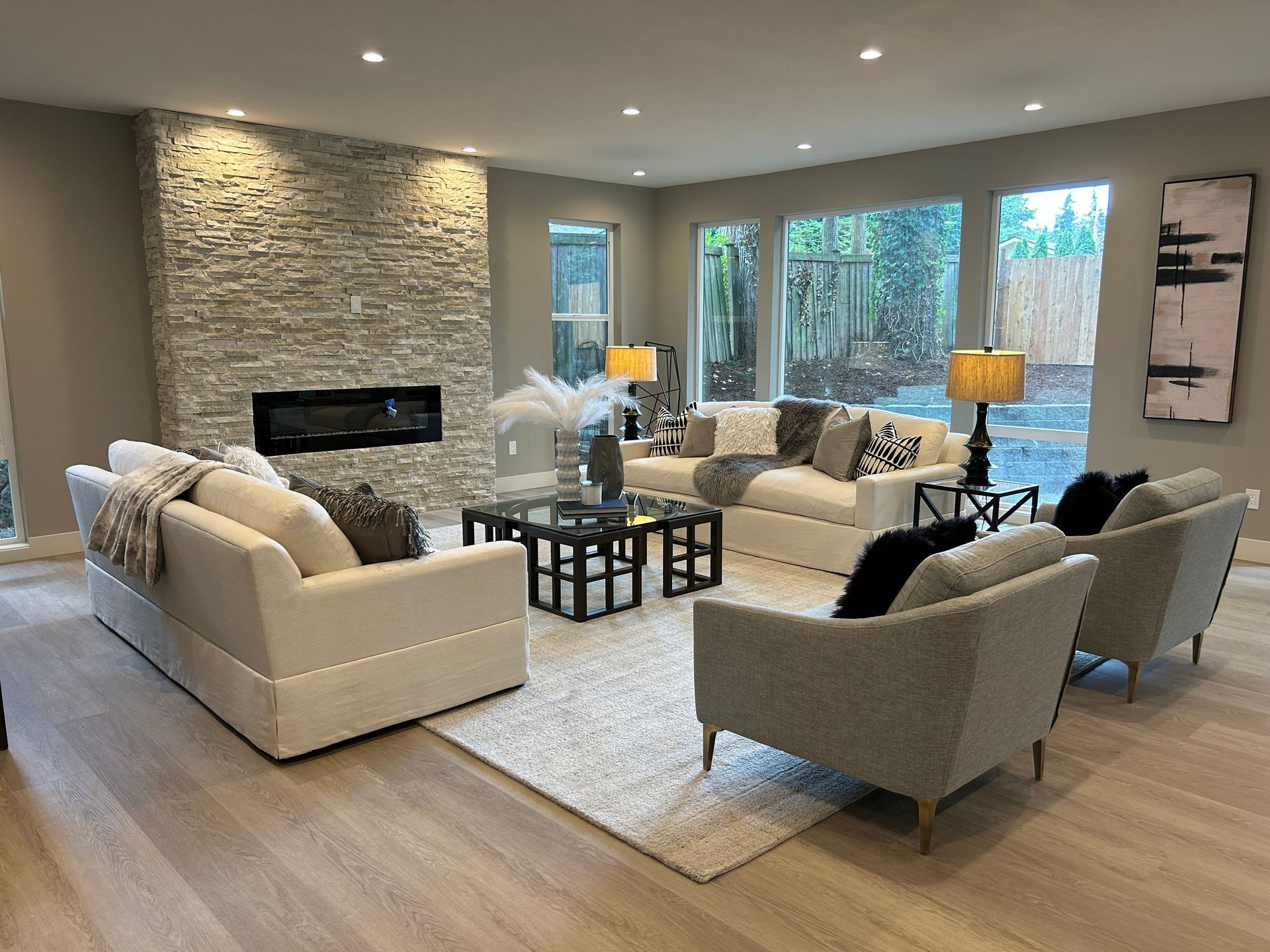 Living room with neutral decor, stone fireplace, white couches, and large windows overlooking greenery.