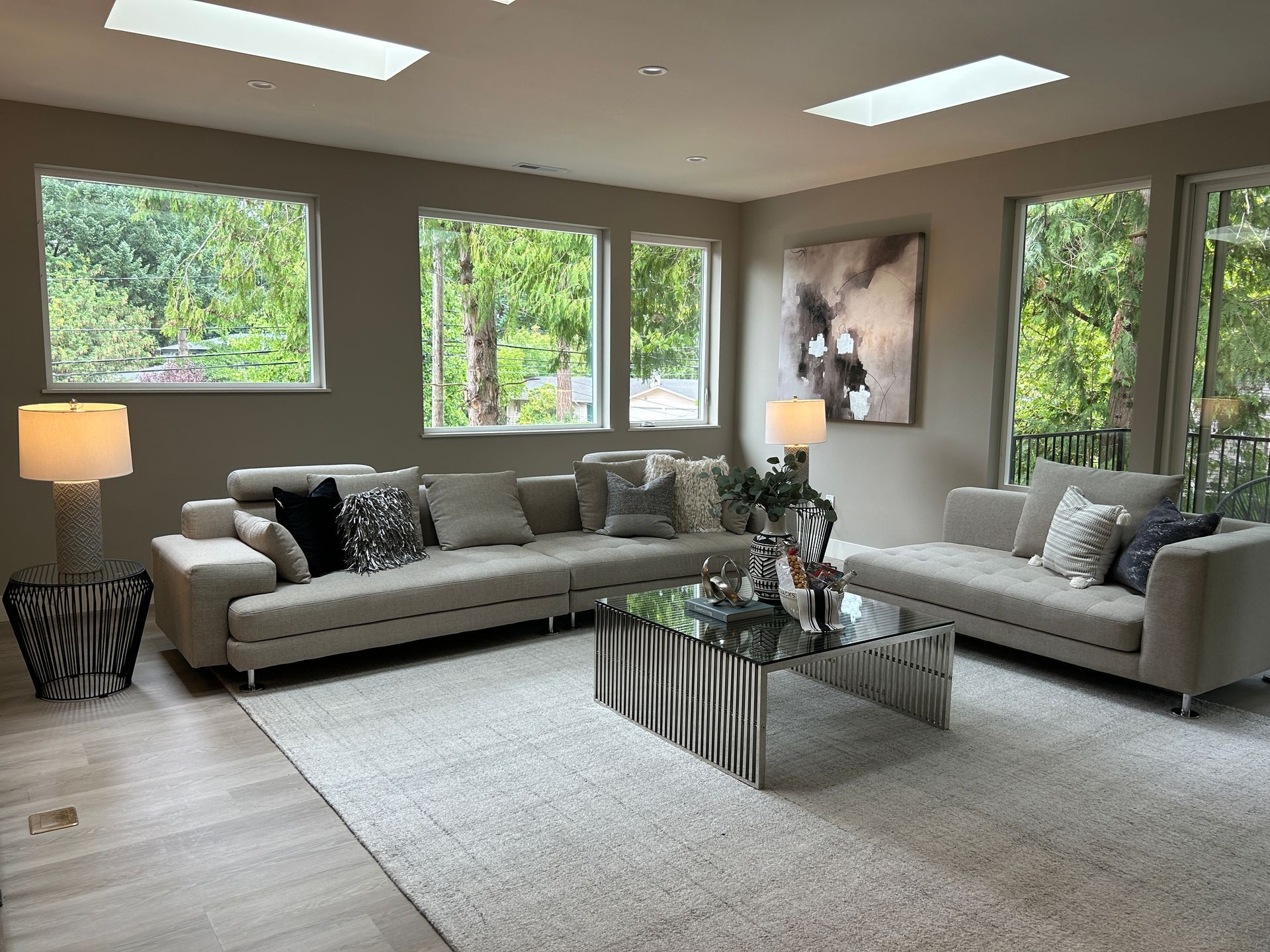 Modern living room with beige couches, mirrored coffee table, large windows with greenery views.