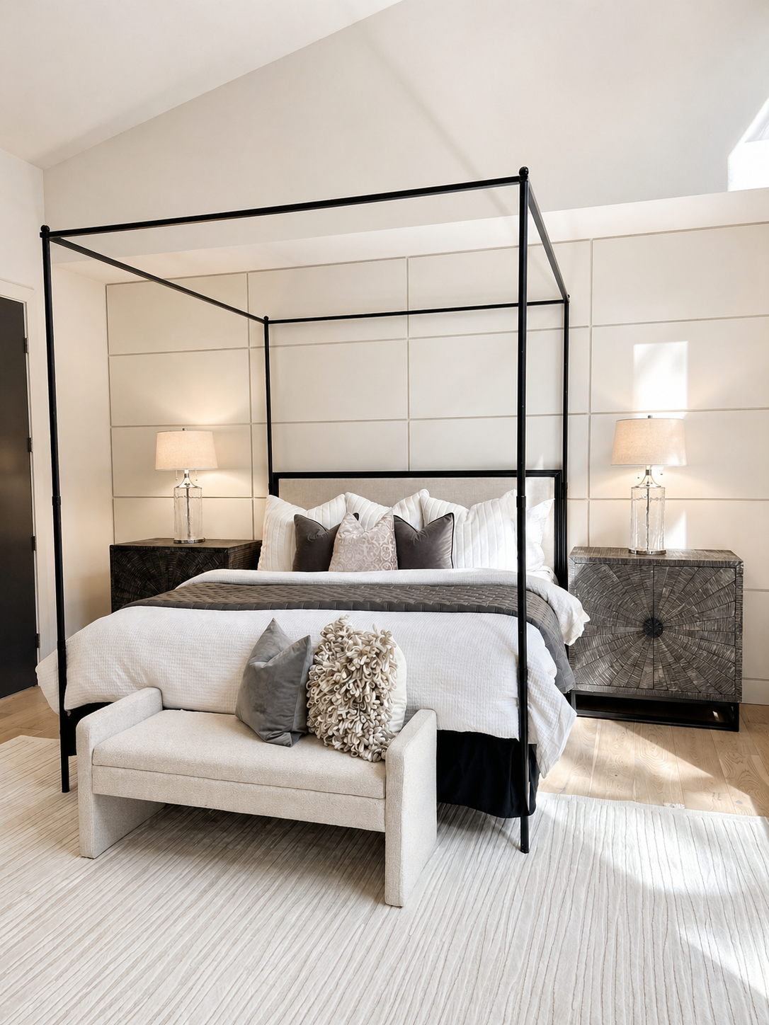Black four-poster bed in a bright bedroom with cream bedding and a tufted bench at the foot.