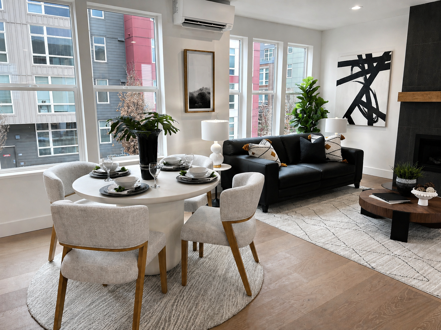 Modern living-dining room with large windows, white table, gray chairs, black sofa, fireplace, and city view.