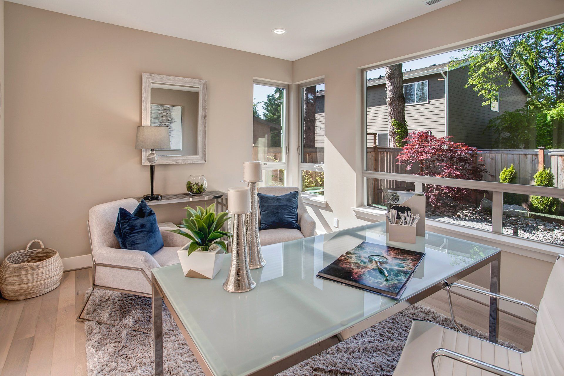 Home office with desk, large windows, white chairs, and blue pillows.