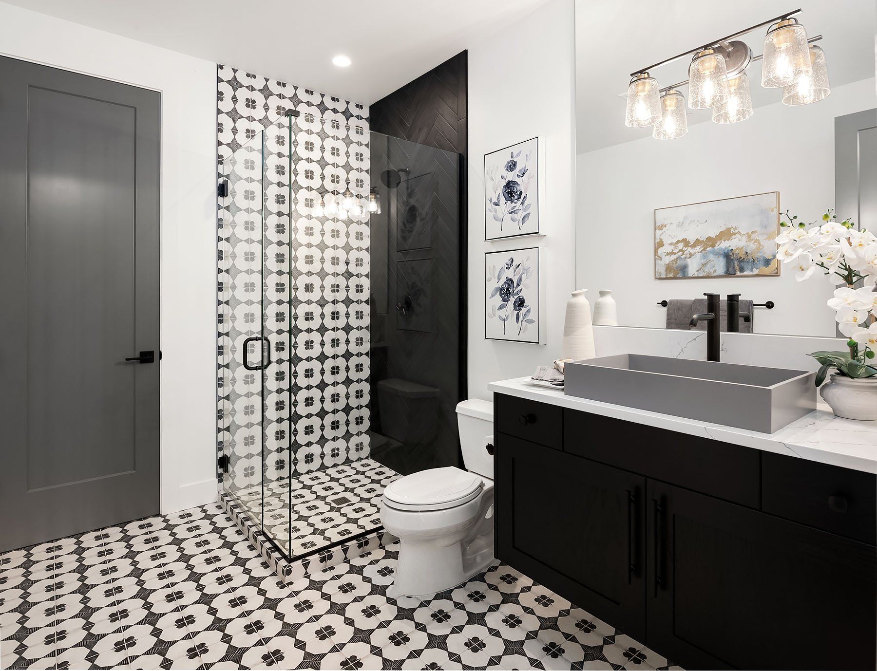 Modern bathroom: patterned tile floor, black vanity, glass shower, gray door.