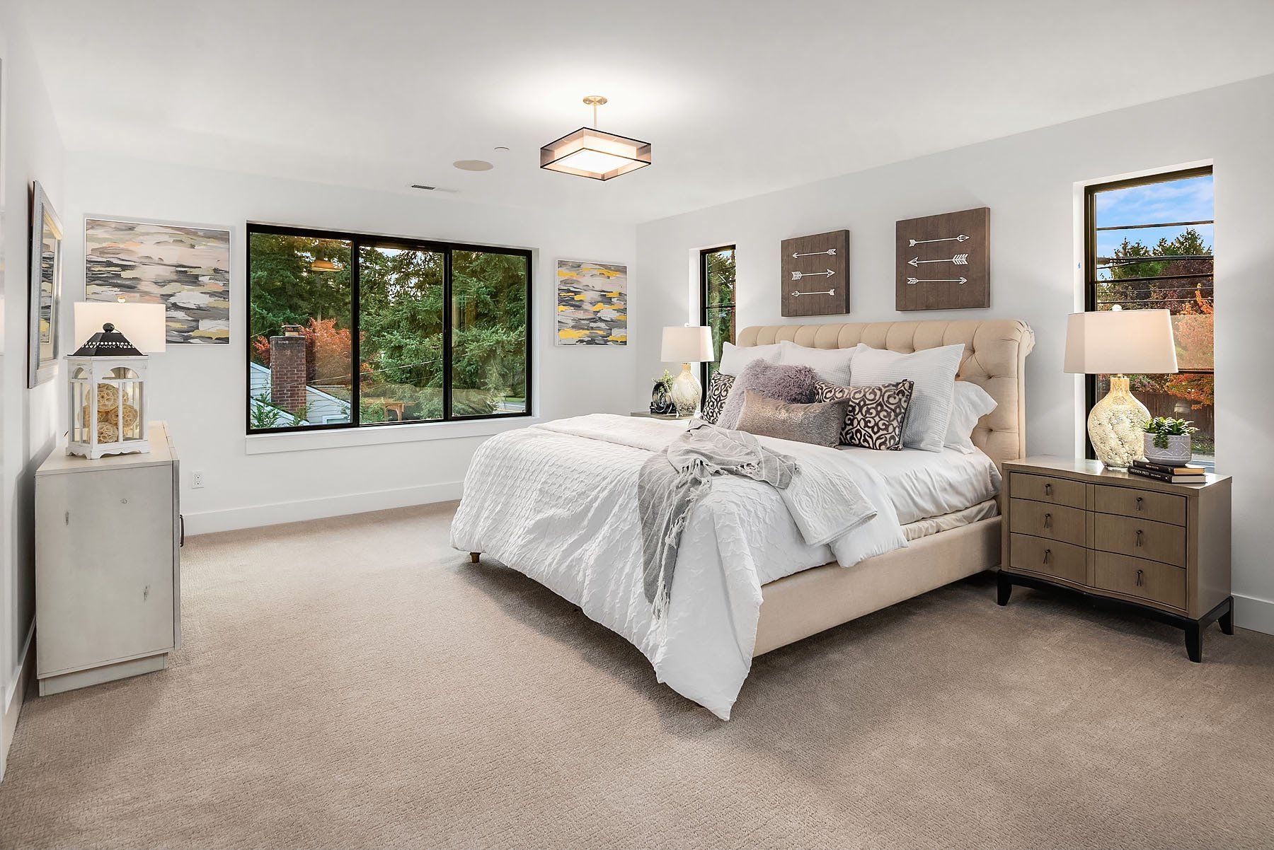 Bedroom with large windows, bed, nightstands, art, and light-colored carpet.