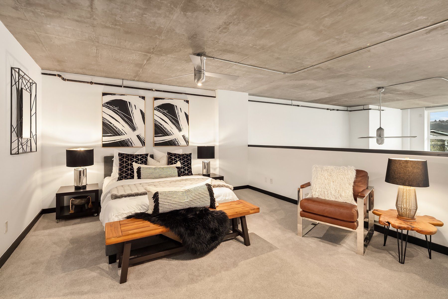 Loft bedroom with exposed concrete ceiling, bed, chair, side tables. Black, white, and brown color scheme.