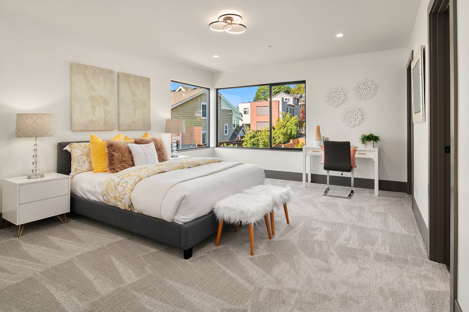 Bedroom with bed, desk, and window; neutral tones with pops of yellow; carpet flooring.