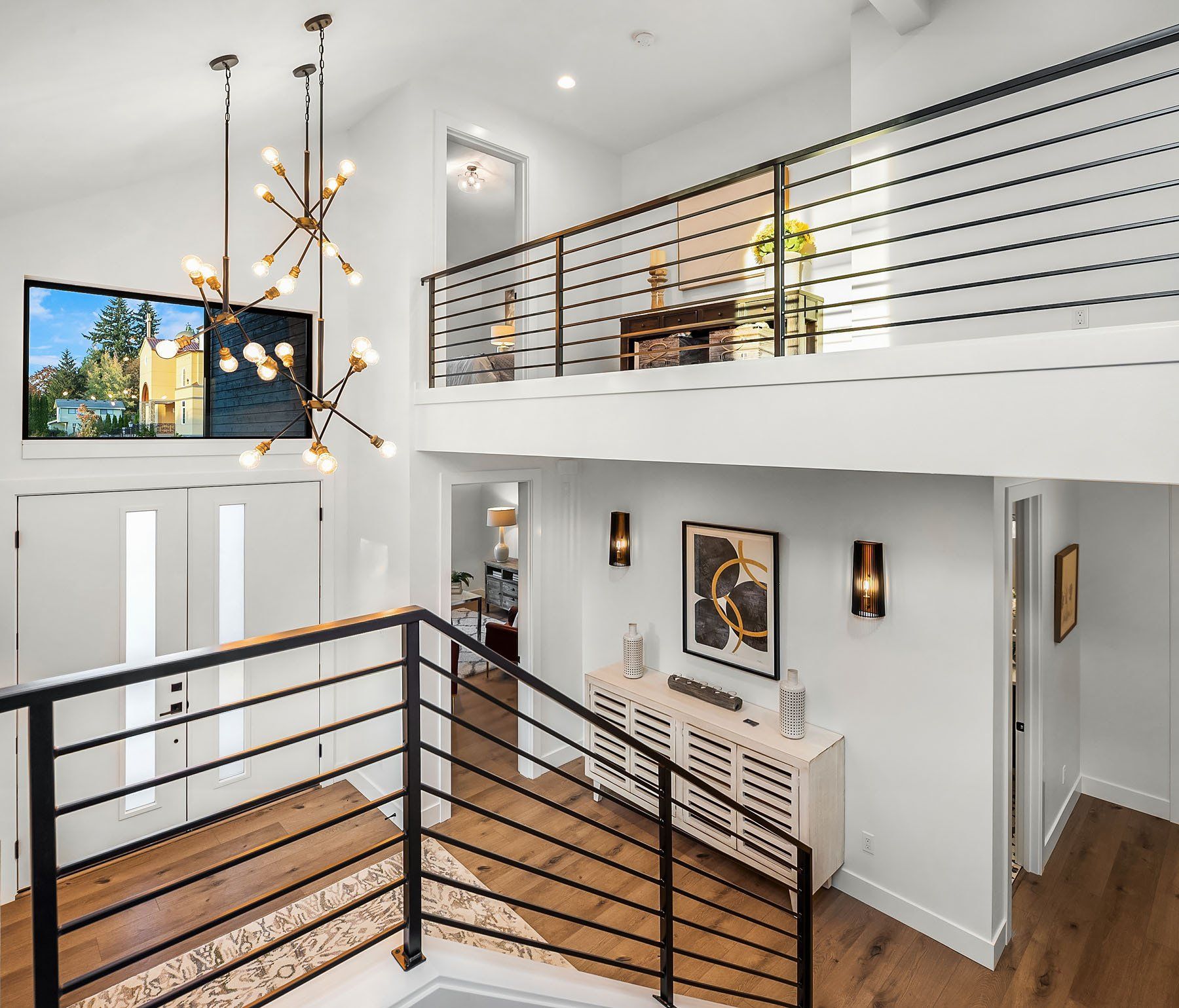 Modern, two-story entryway with wood floors, black railings, and white walls. A large chandelier hangs from the high ceiling.
