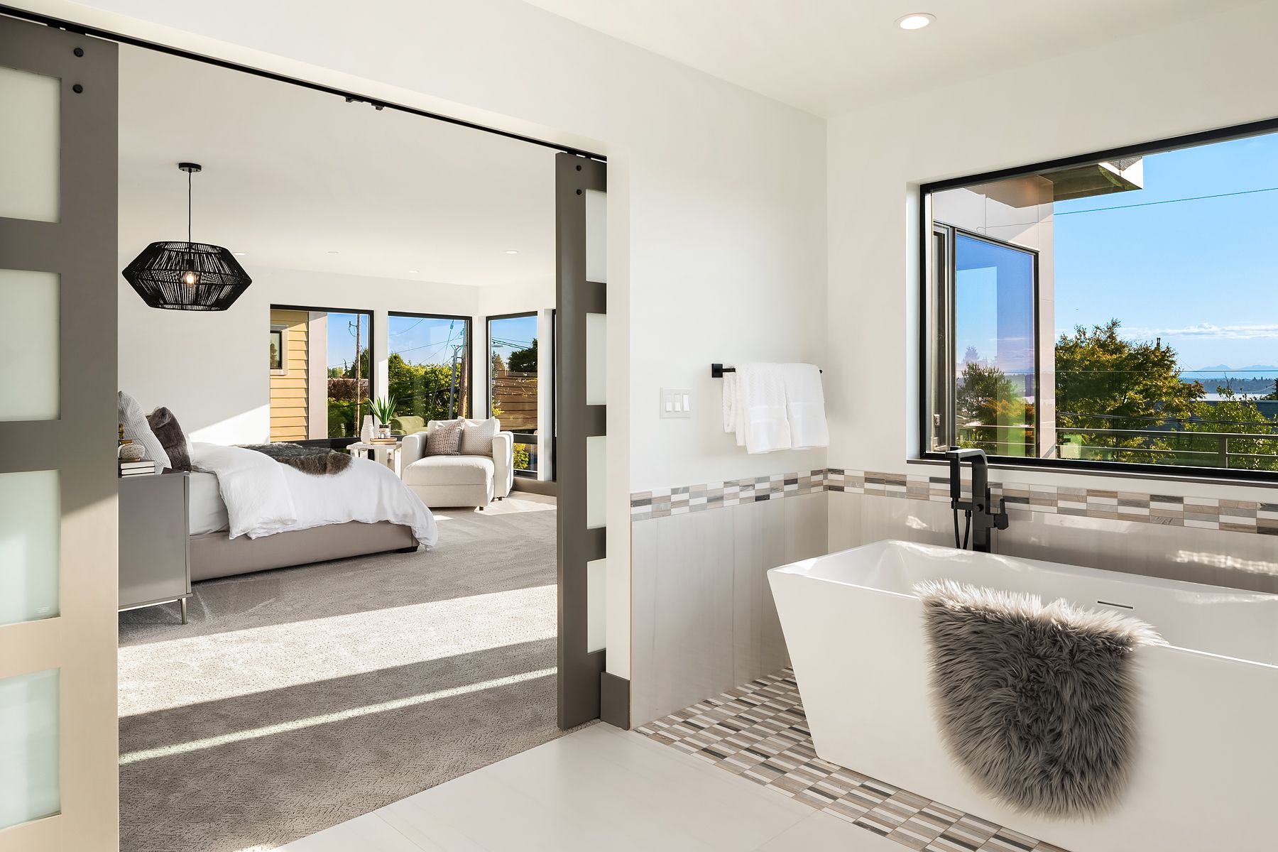 Open bathroom with tub, pebble floor, and open door to bedroom with sunny view.