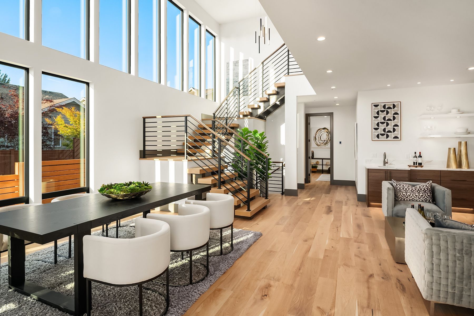 Modern living room with dining area, staircase, and large windows overlooking fall foliage.
