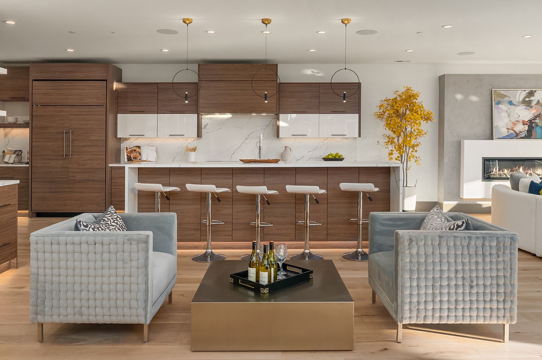 Modern kitchen and living room with wooden cabinets, white countertops, and two gray armchairs facing a gold coffee table.