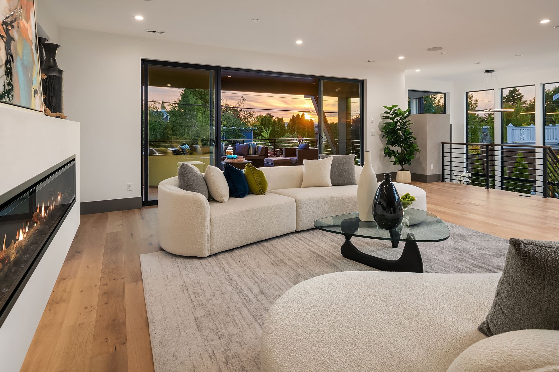 Modern living room with fireplace, plush sofa, and large windows overlooking a sunset.