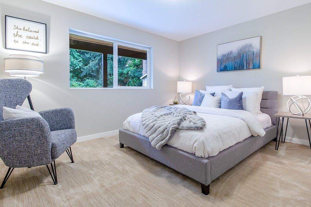 Cozy bedroom with a bed, armchair, and a window overlooking trees. Neutral tones, soft lighting.