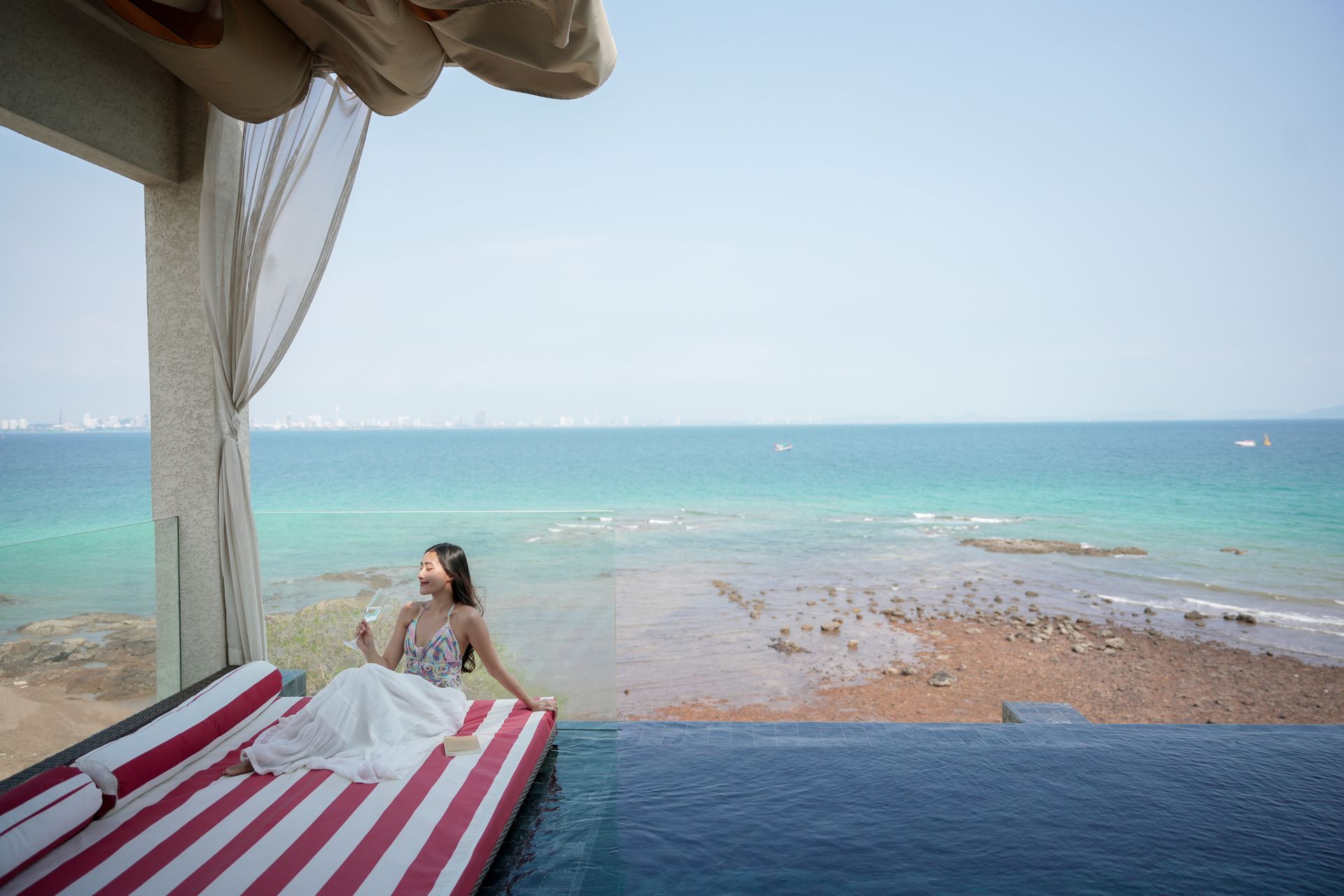 Woman in white dress reclines on striped lounge by infinity pool overlooking ocean.