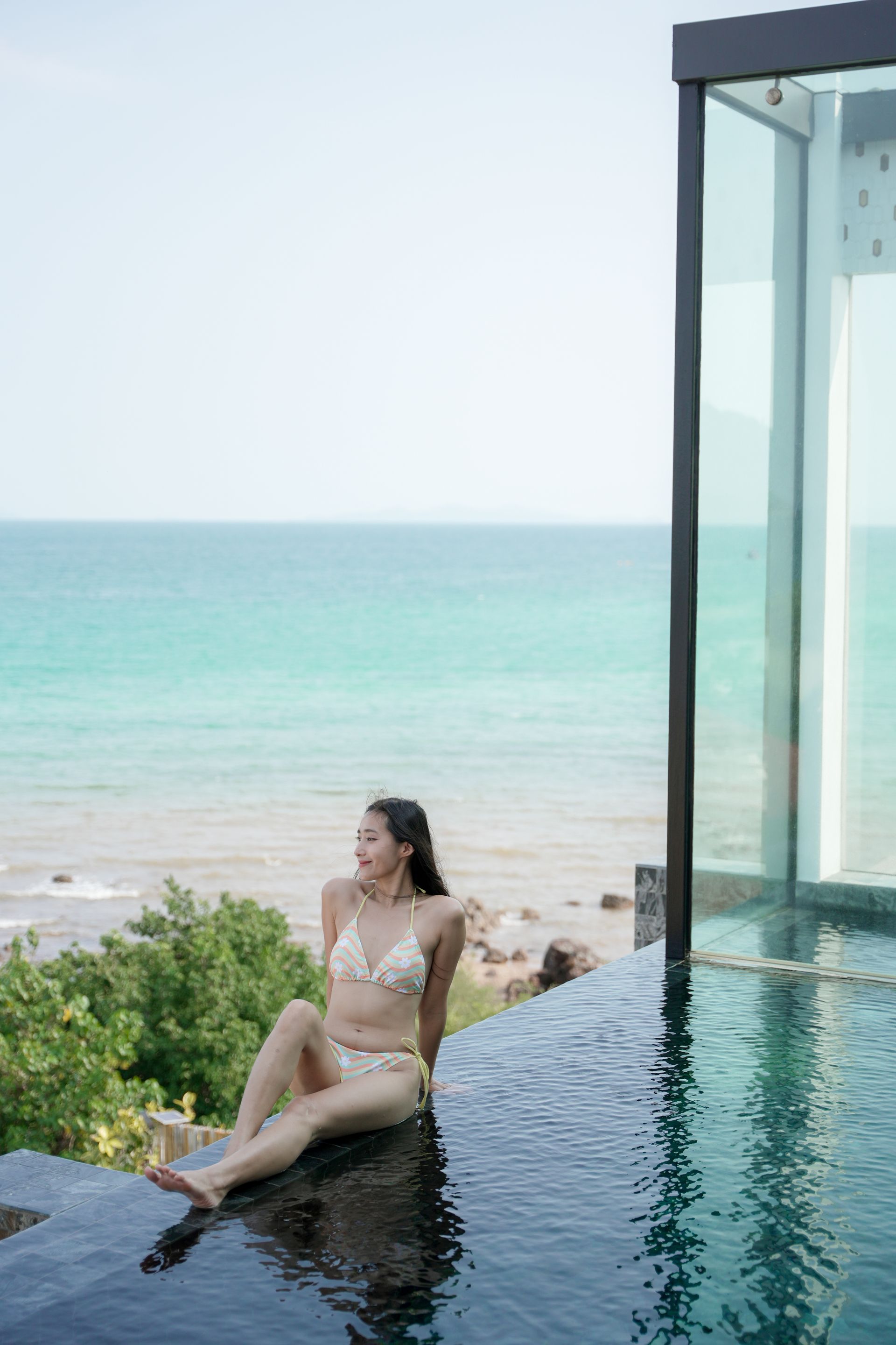 Woman in bikini sits by pool, looking at ocean view.