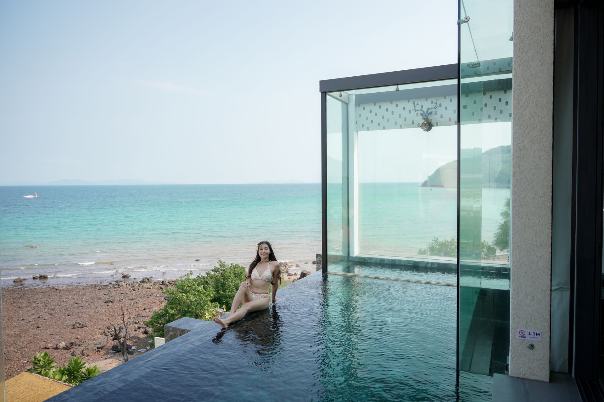 Woman in swimsuit relaxing by an infinity pool overlooking the ocean. Glass walls, sunny day.