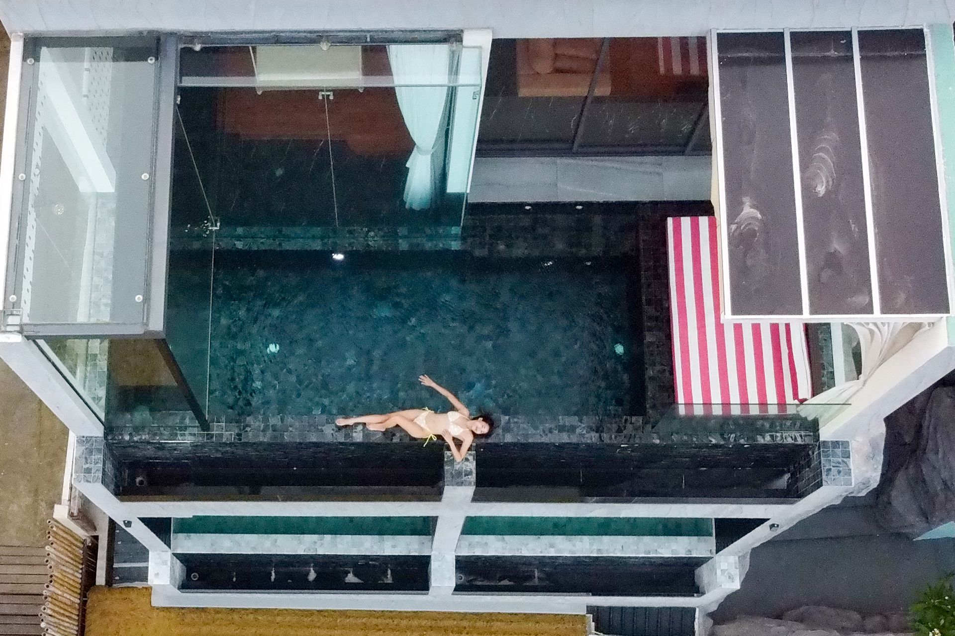 Overhead view of a person relaxing by a dark-tiled pool with a glass enclosure; a striped towel rests nearby.