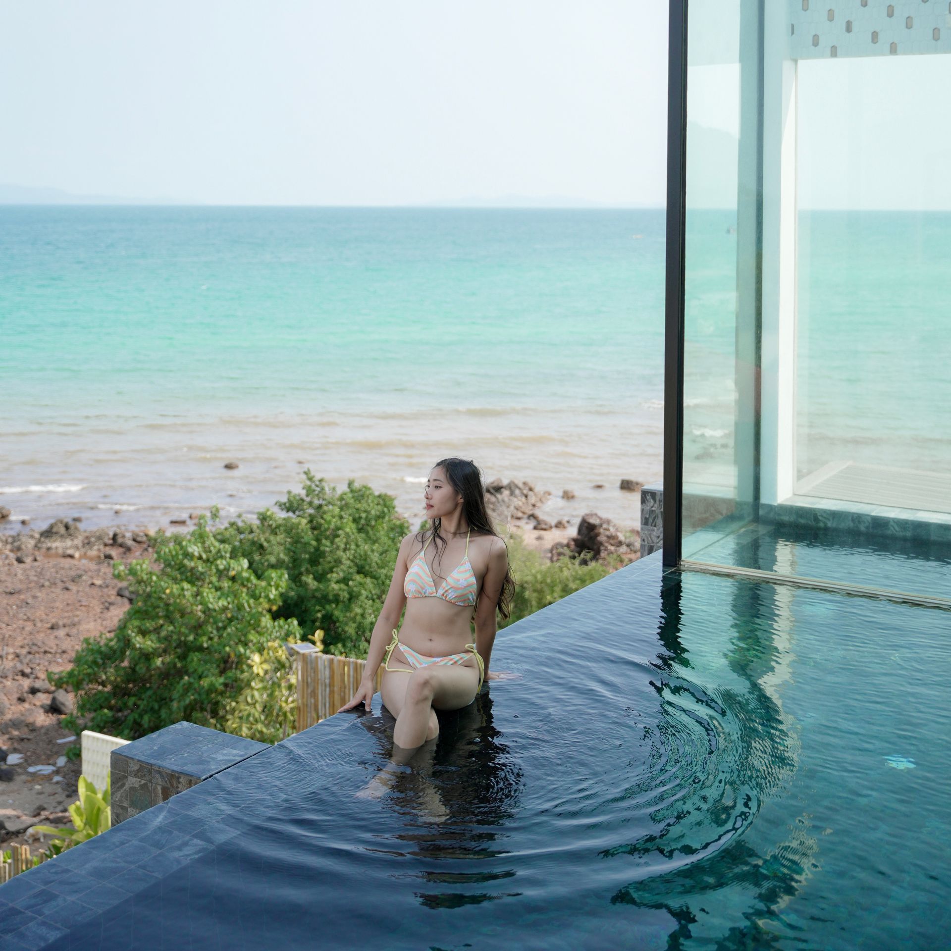 Woman sits on the edge of an infinity pool, looking out at the ocean.