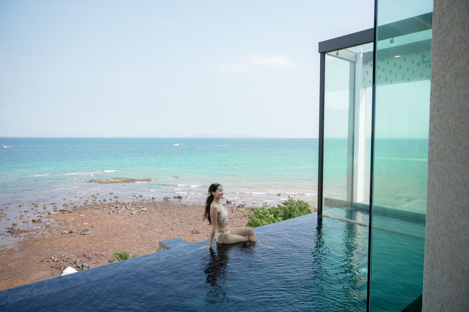 Woman in a swimsuit sits at the edge of an infinity pool overlooking the ocean.