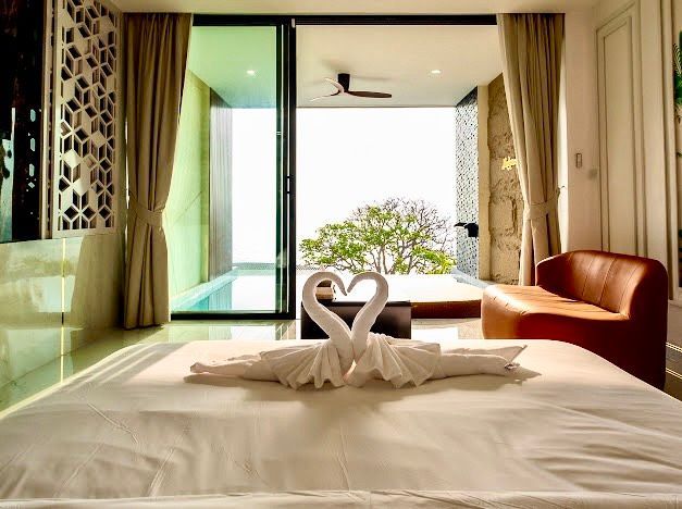 Hotel room interior: bed with towel swans, sliding door to pool and trees, brown couch.