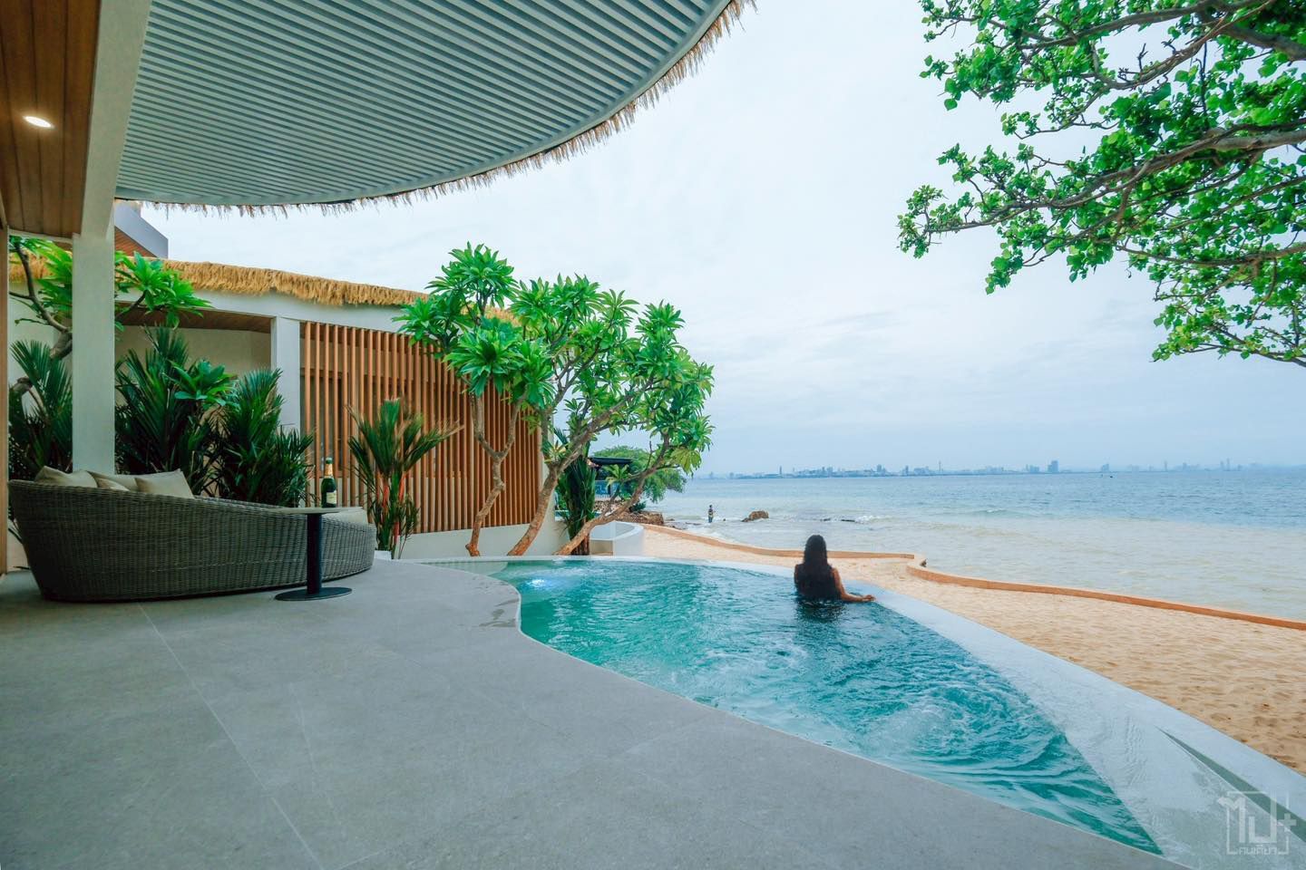 Infinity pool next to beach. Person in pool, ocean view. Outdoor seating area with wooden accents. Cloudy sky.