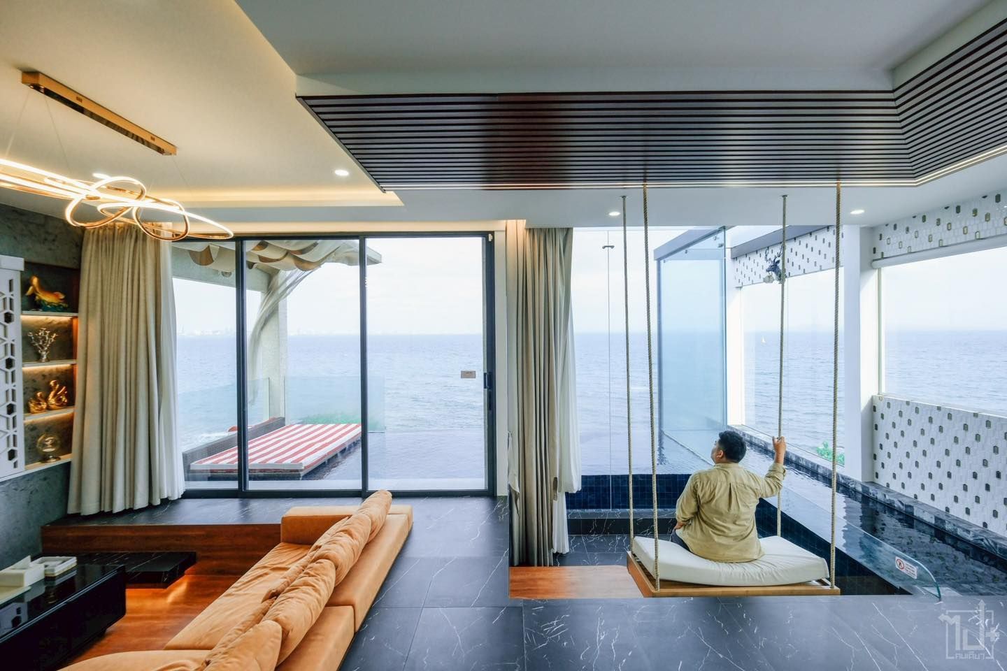 Living room with ocean view, person on swing, tan sofa, open doors, and decorative ceiling.